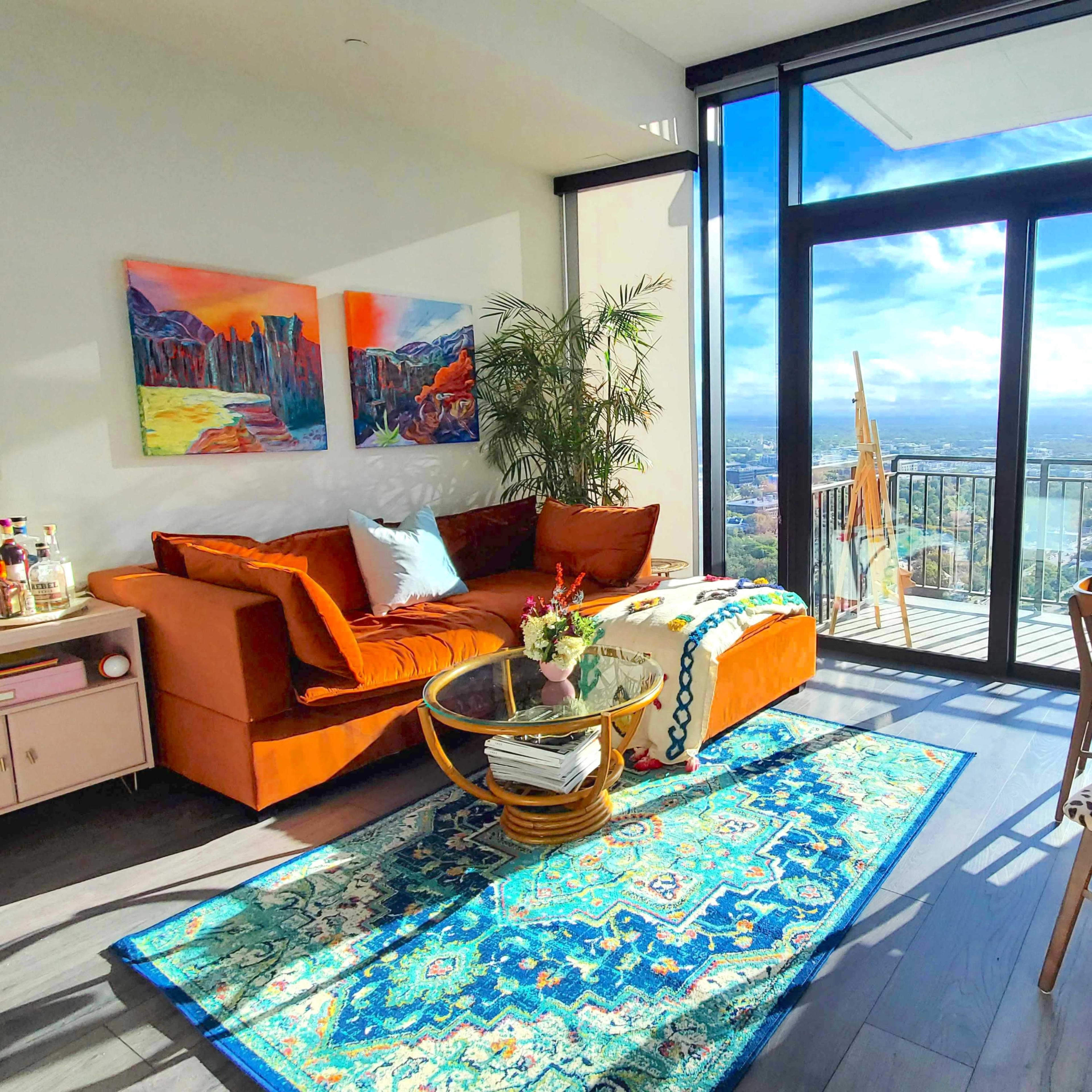 A modern living room features a brown sofa, colorful artwork on the walls, a glass coffee table, and a view of the city through large windows.