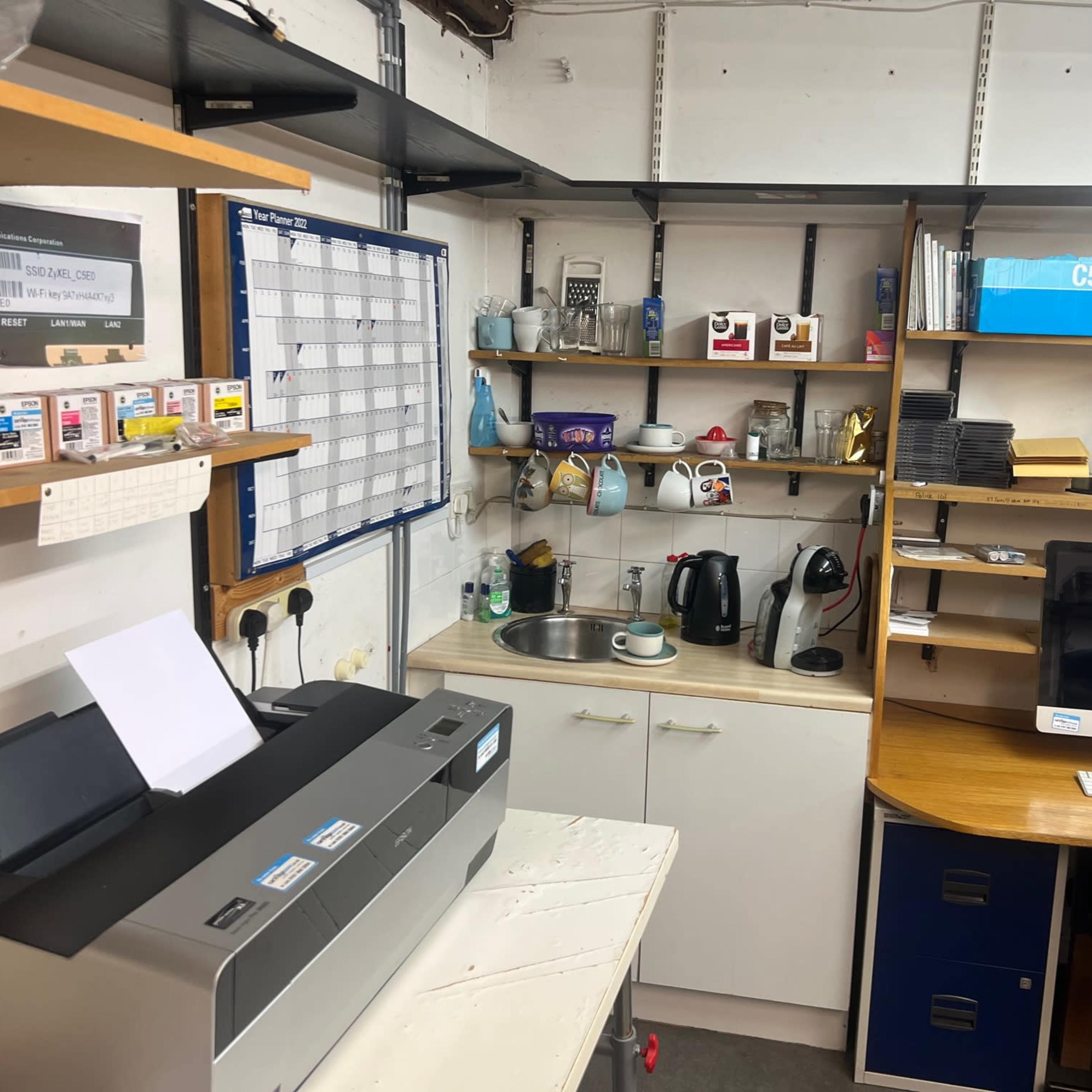 A small workspace featuring a printer, a calendar on the wall, shelves with various items, a sink, kitchen appliances, and a computer setup.