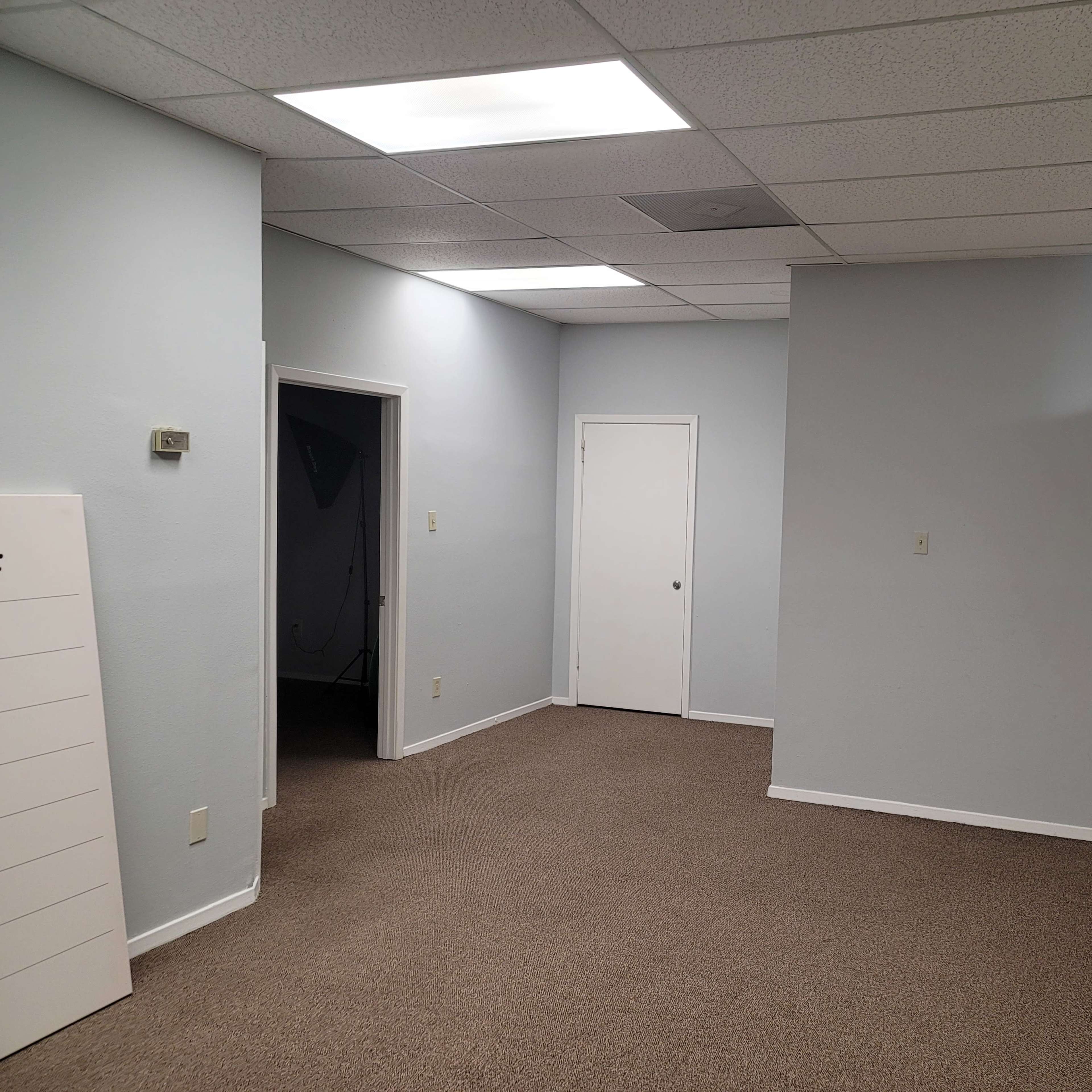 A vacant office space with light gray walls, a carpeted floor, and an interior door on one side.