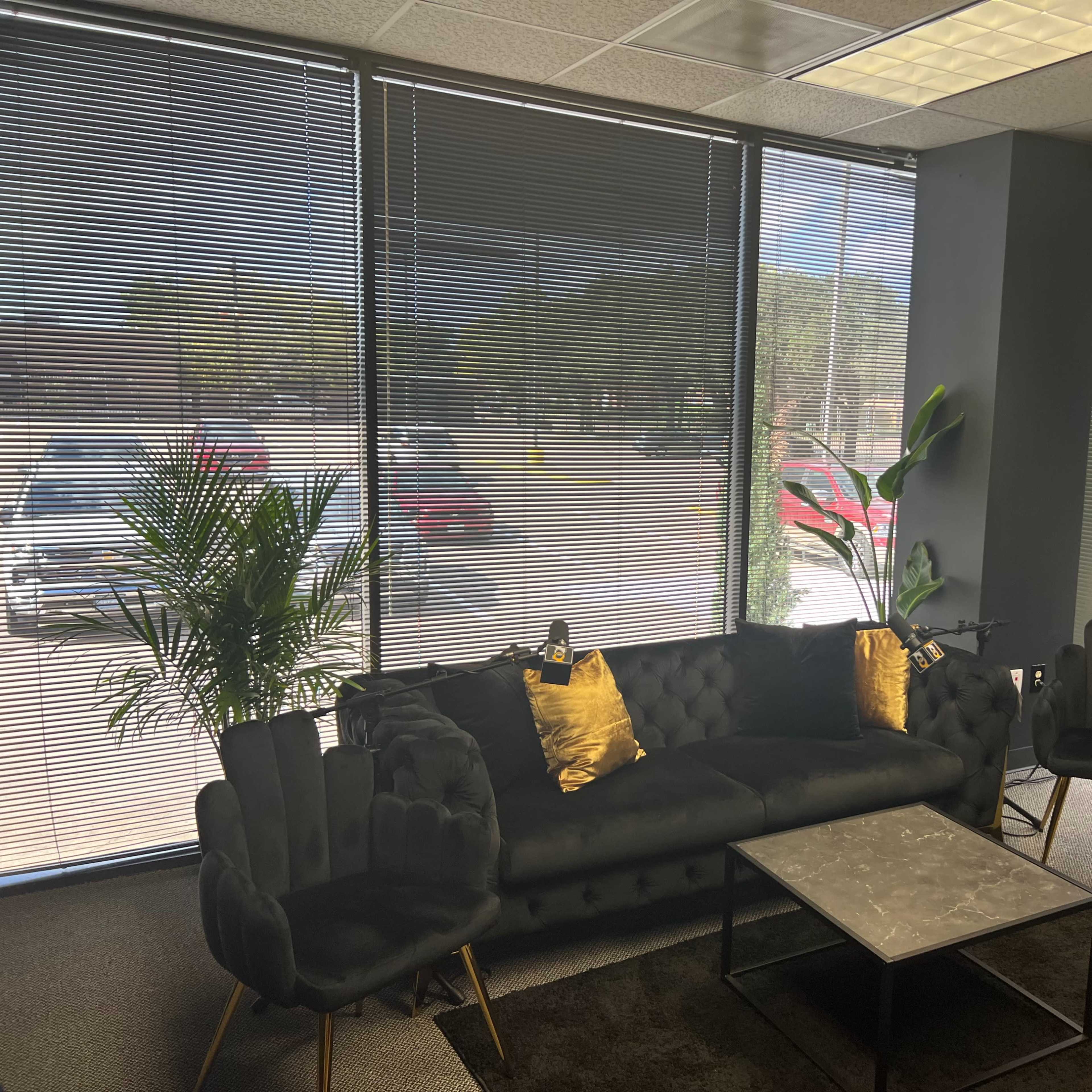 A modern office lounge area featuring a black sofa with gold throw pillows, two matching armchairs, a coffee table, and large windows with blinds allowing natural light.