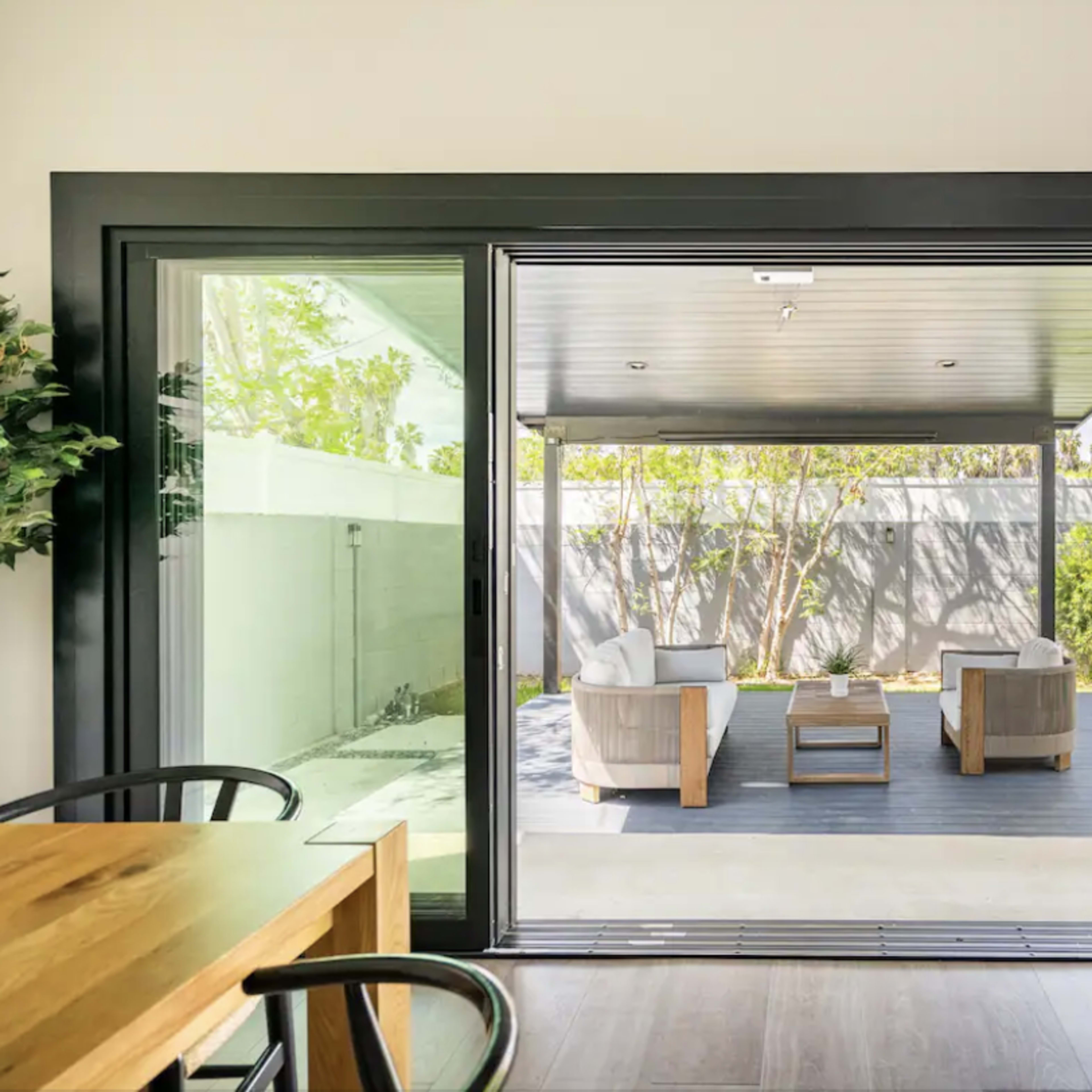 A modern indoor space opens to a patio featuring two armchairs, a coffee table, and a landscaped yard.