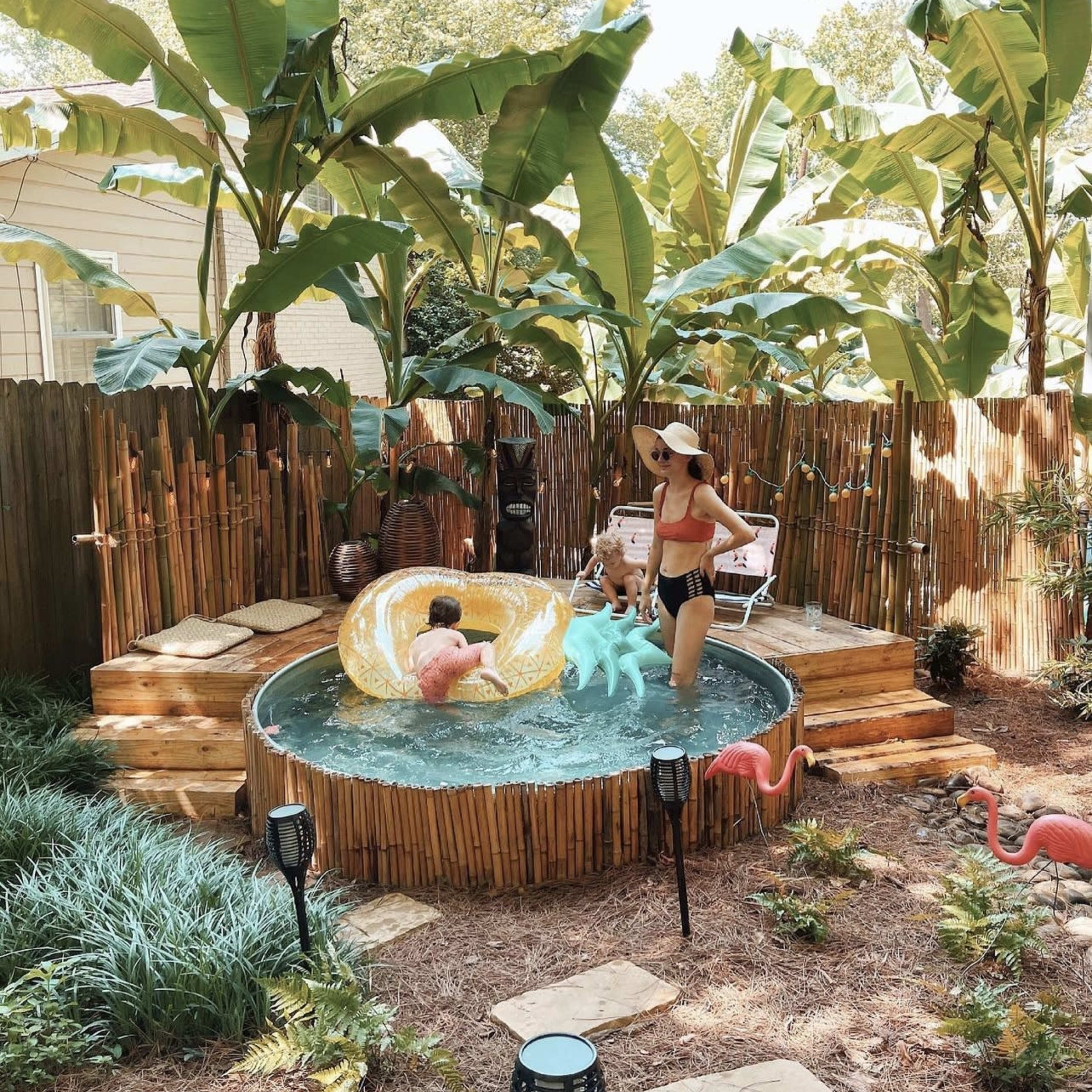 A boy plays in a small pool while a woman stands nearby in a backyard surrounded by tall plants and decorative flamingos.