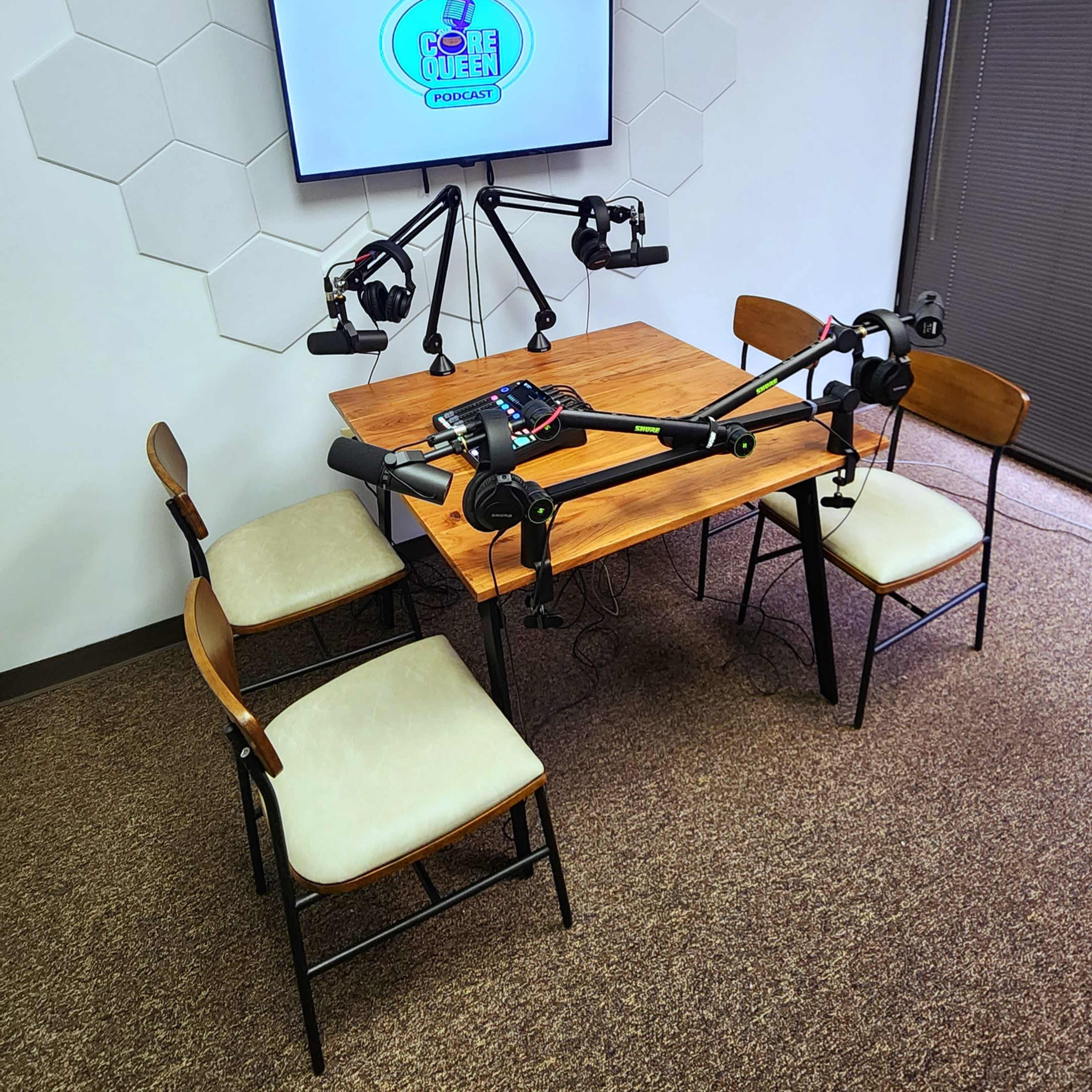 A small podcast recording setup featuring a wooden table with four chairs, equipped with microphone arms and a monitor displaying a podcast logo.