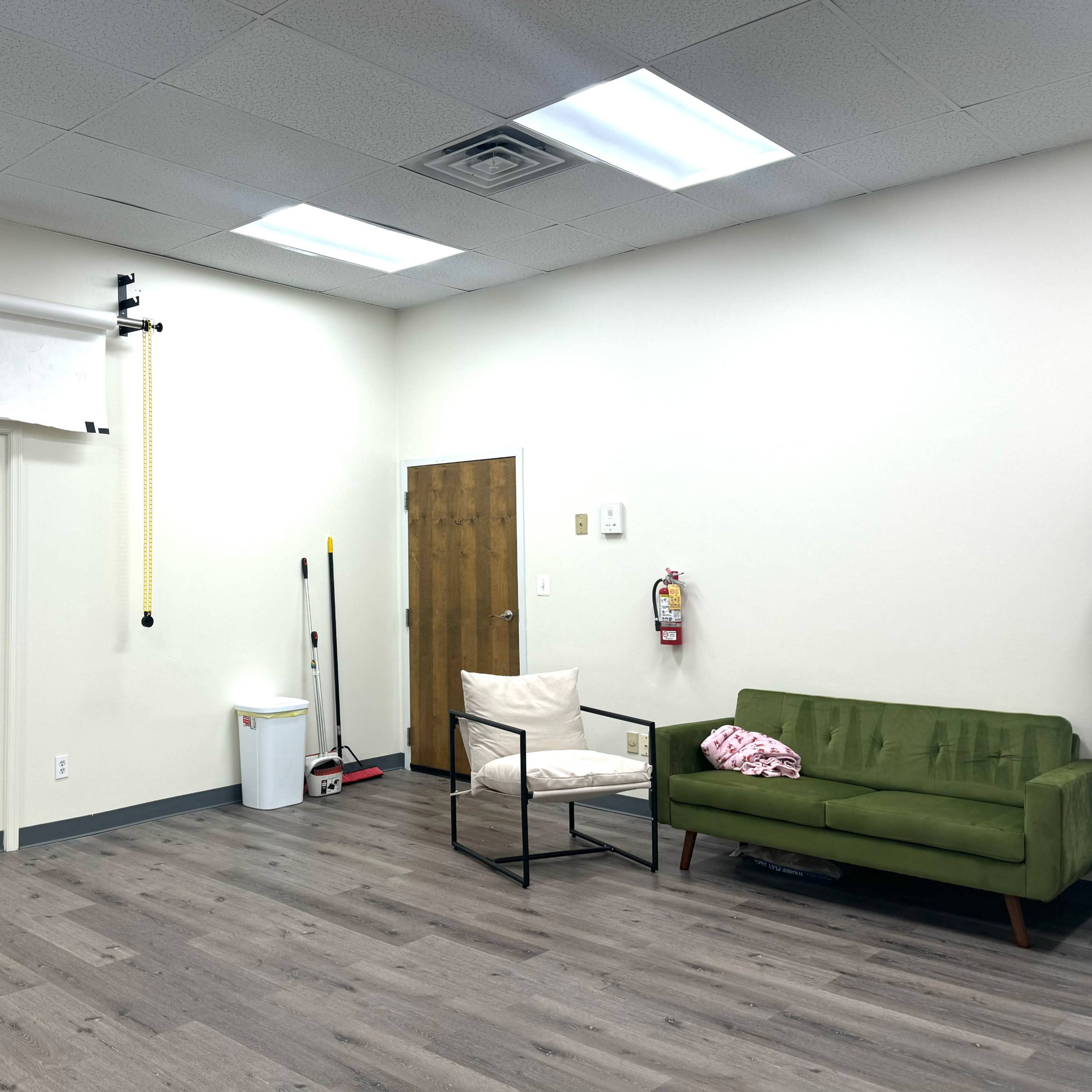A sparse room featuring a green sofa, a black armchair, a wastebasket, and a door on one wall, with a roll of backdrop paper mounted above a plain wall.