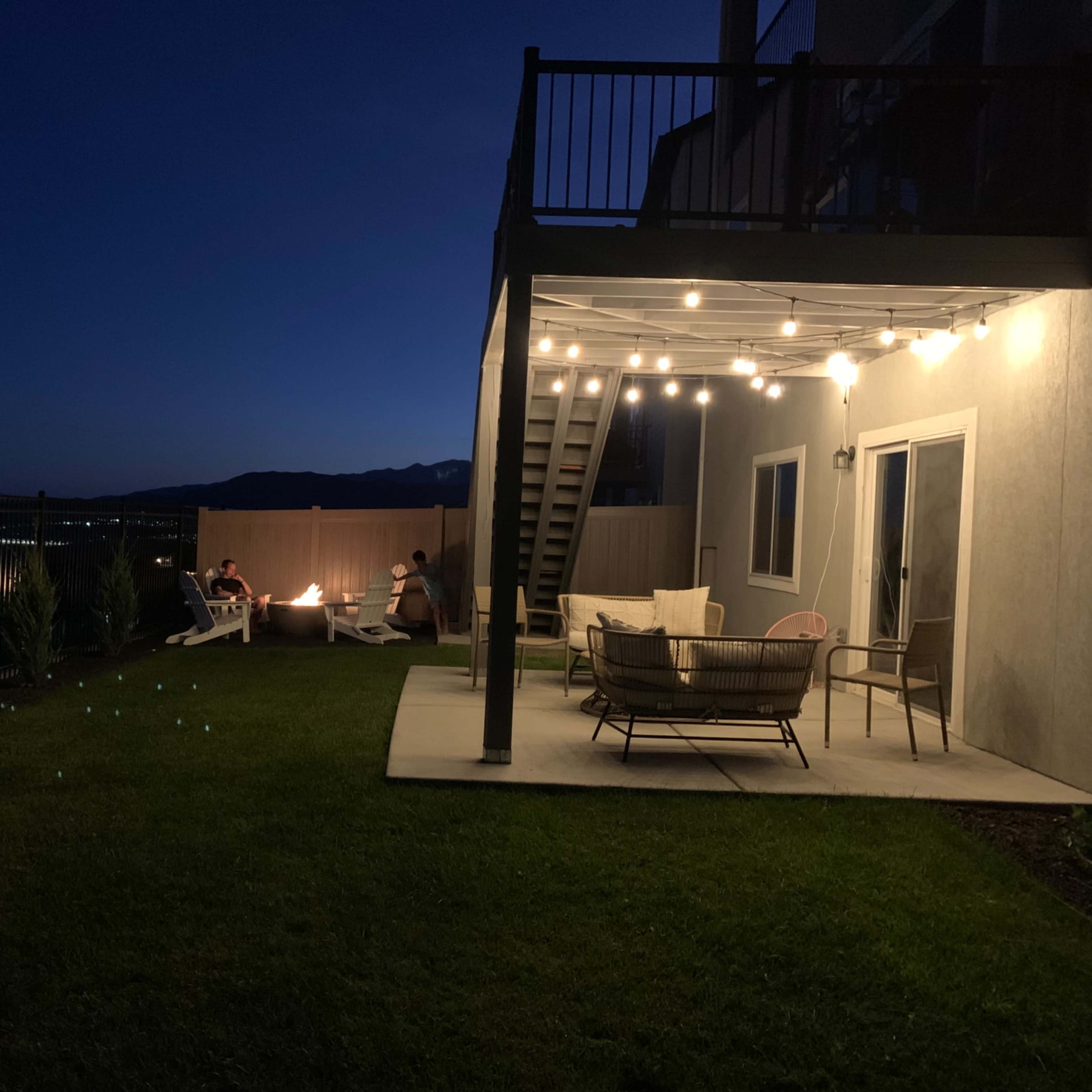 A backyard at night features two people sitting around a fire pit, with decorative string lights illuminating a patio area and views of mountains in the distance.
