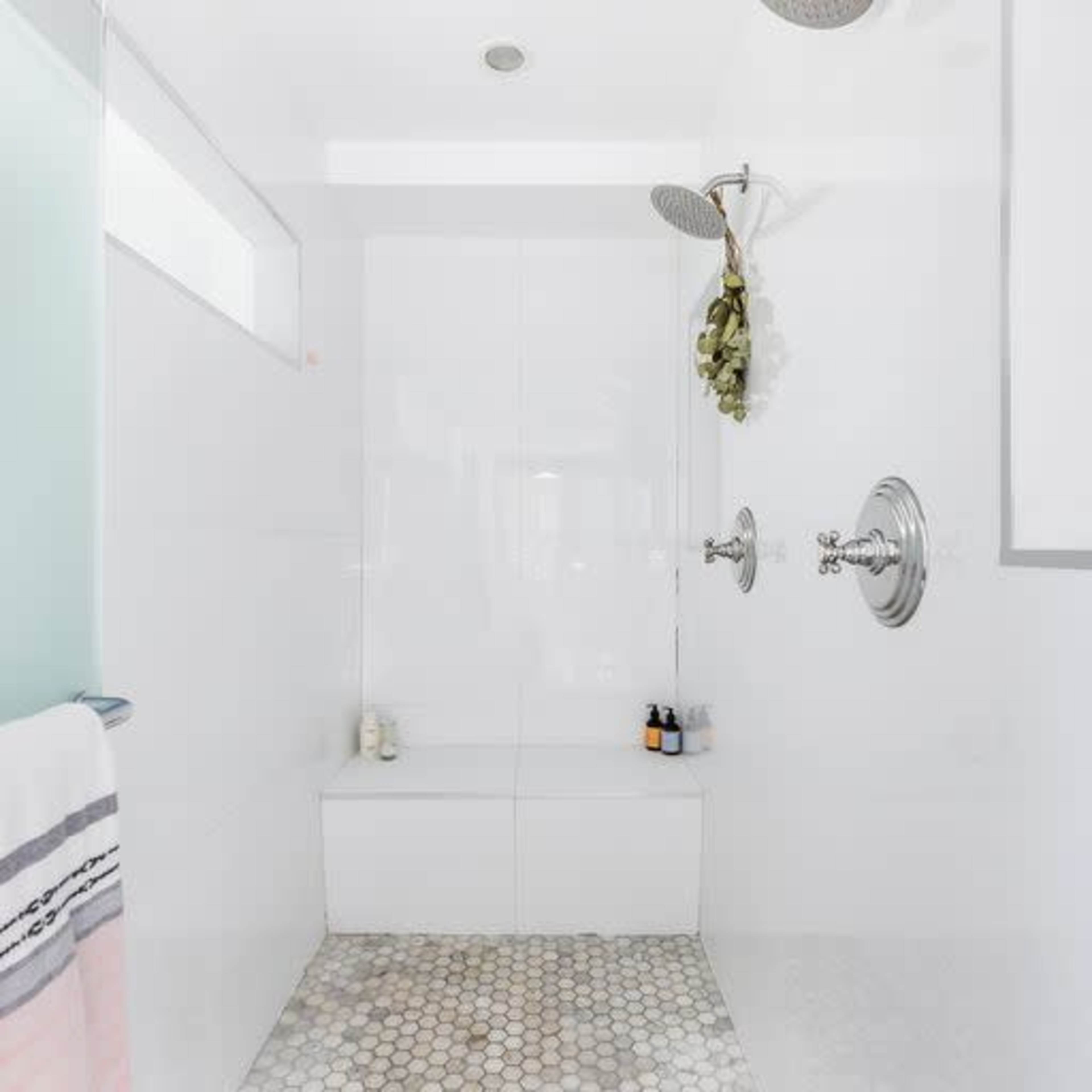 A modern shower area with white tile walls, a showerhead, and a small bench, featuring a pebble stone floor.