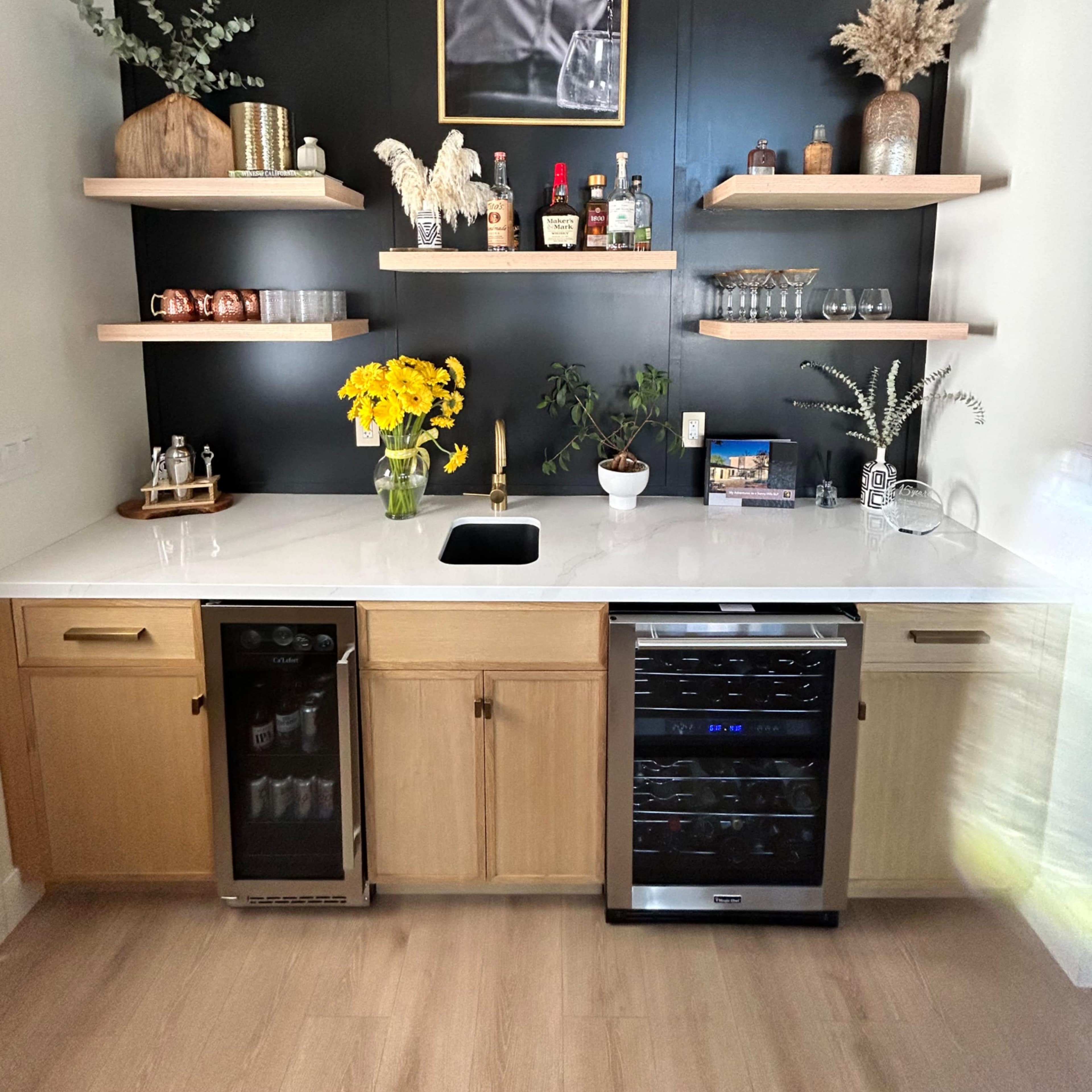 A modern kitchen bar area with wooden cabinets, a black accent wall, open shelves displaying various bottles and decor, and a sink with a countertop adorned with yellow flowers.