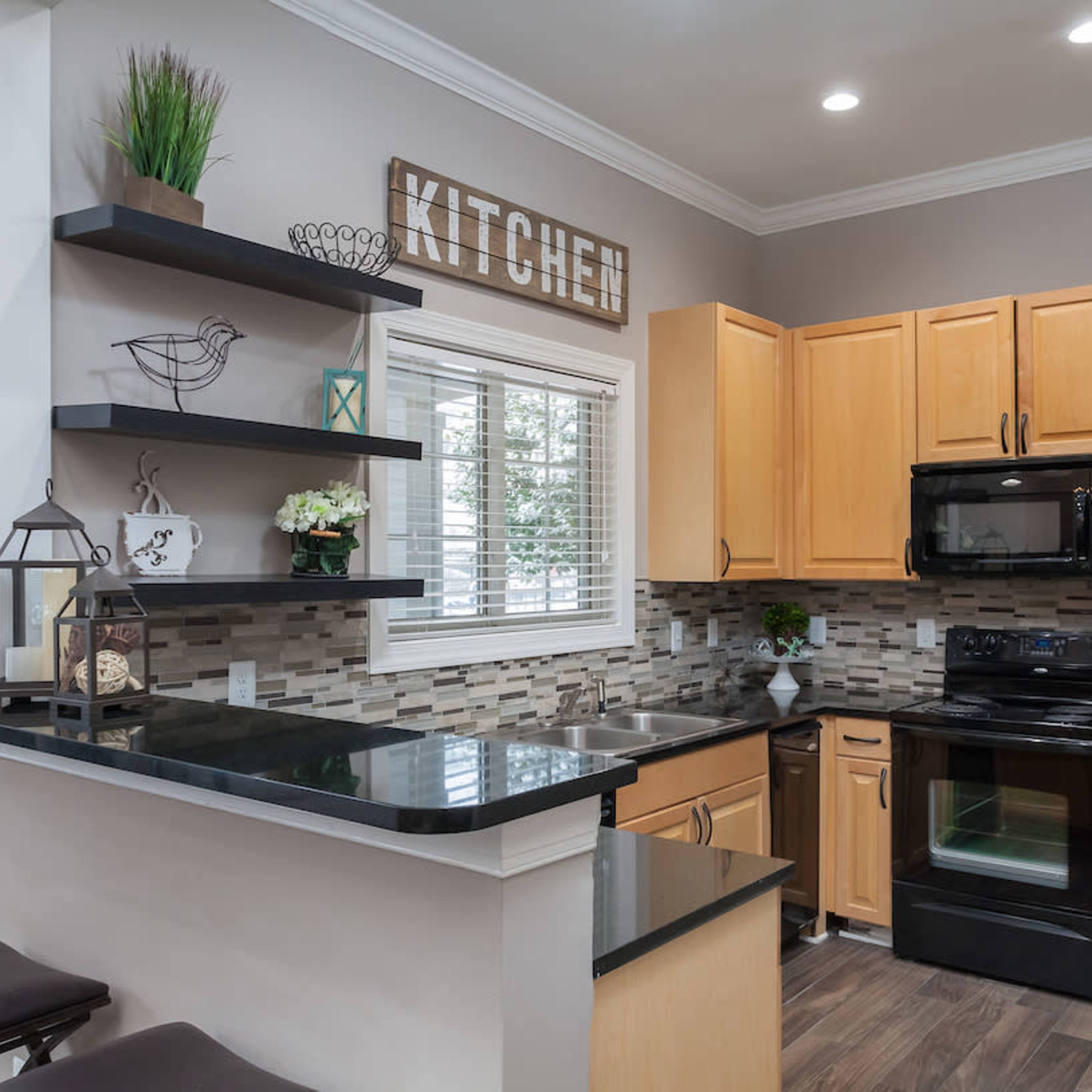 A modern kitchen features wooden cabinets, a black countertop, and stainless steel appliances, with decorative shelves and a window above the sink.