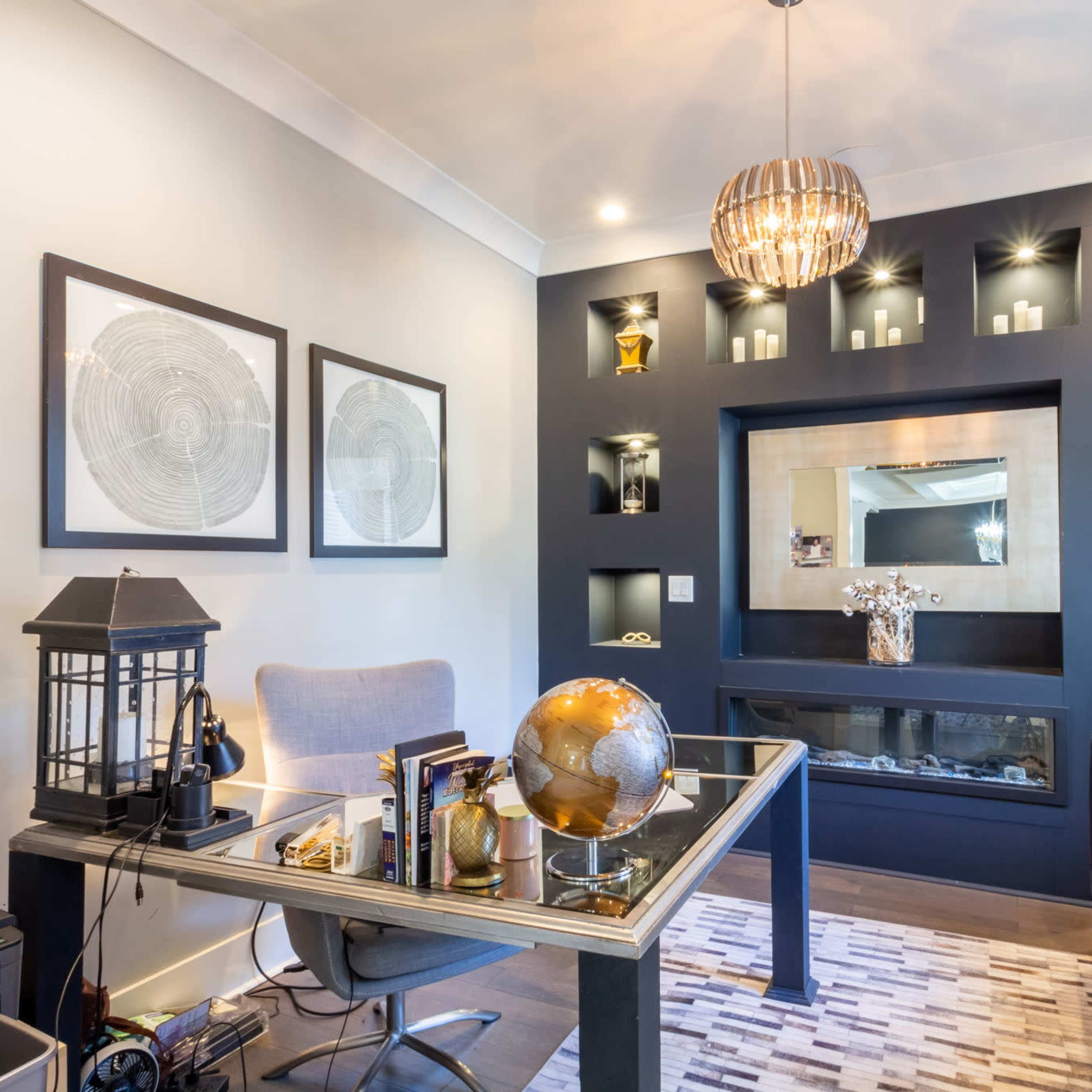 A modern office features a glass-top desk with a globe and decorative items, accented by dark walls with shelves showcasing various objects and framed artwork.