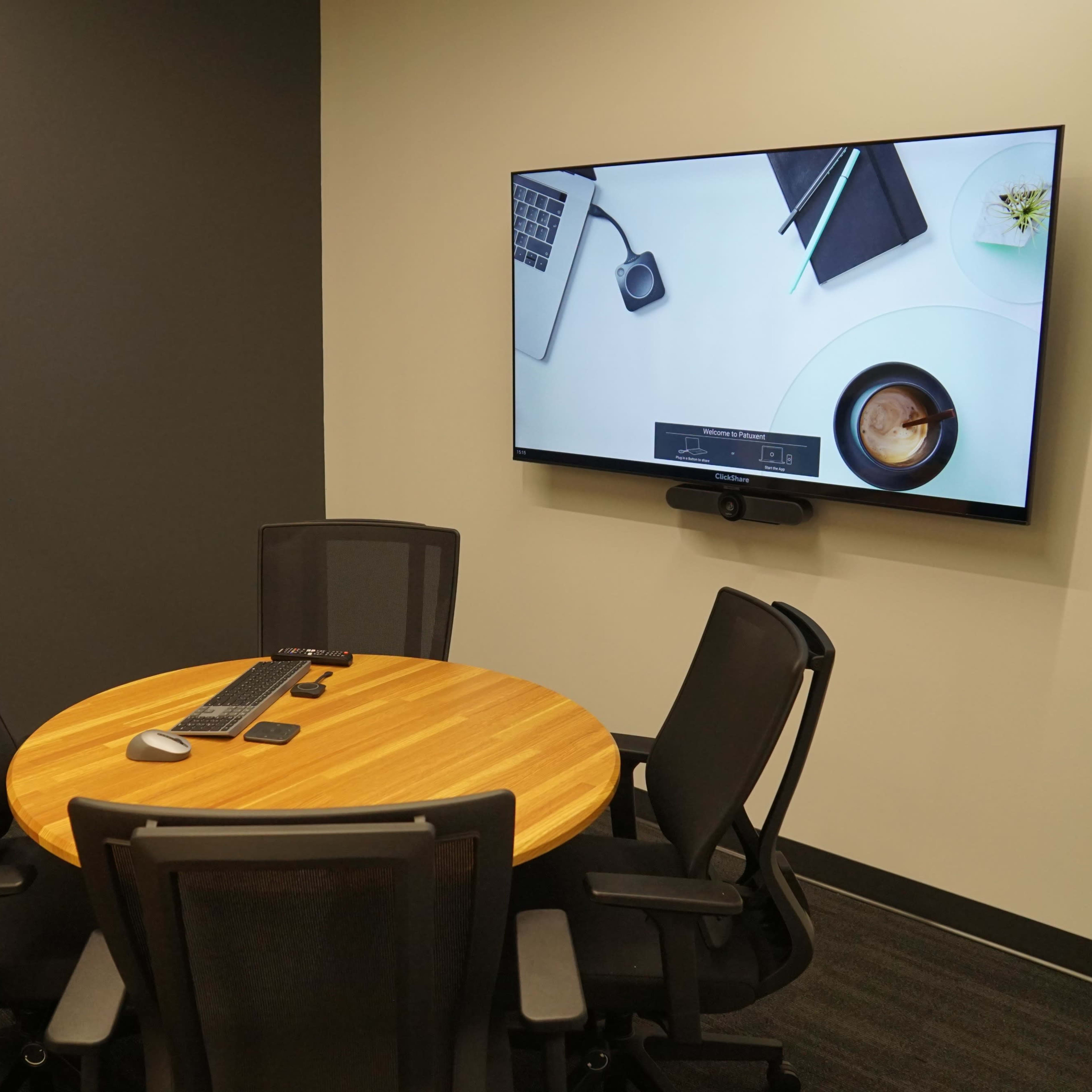 A small conference room features a round wooden table with four black chairs and a large wall-mounted screen displaying a laptop and a cup of coffee.