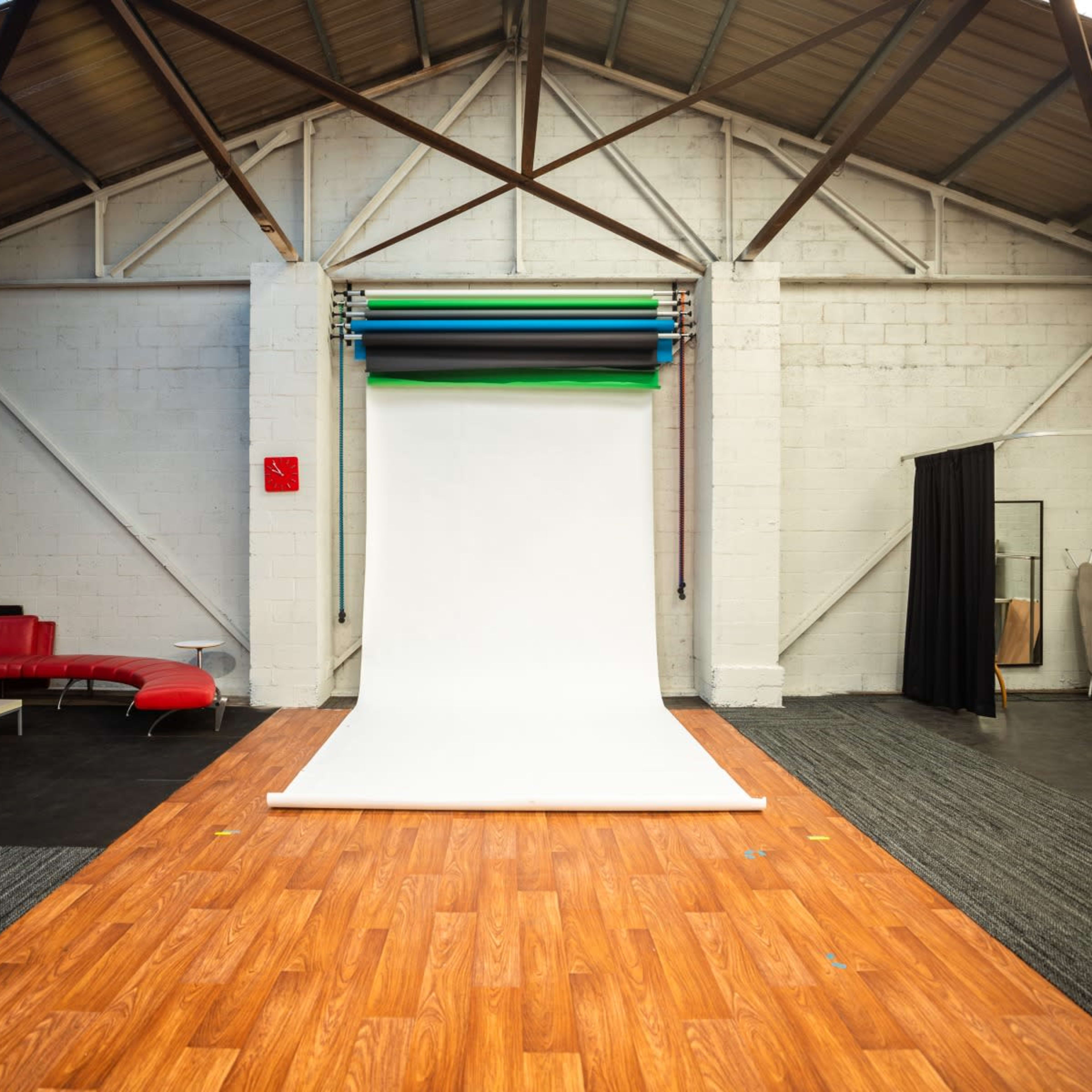 A spacious photography studio with a white backdrop, wooden flooring, a red couch, and black curtains.