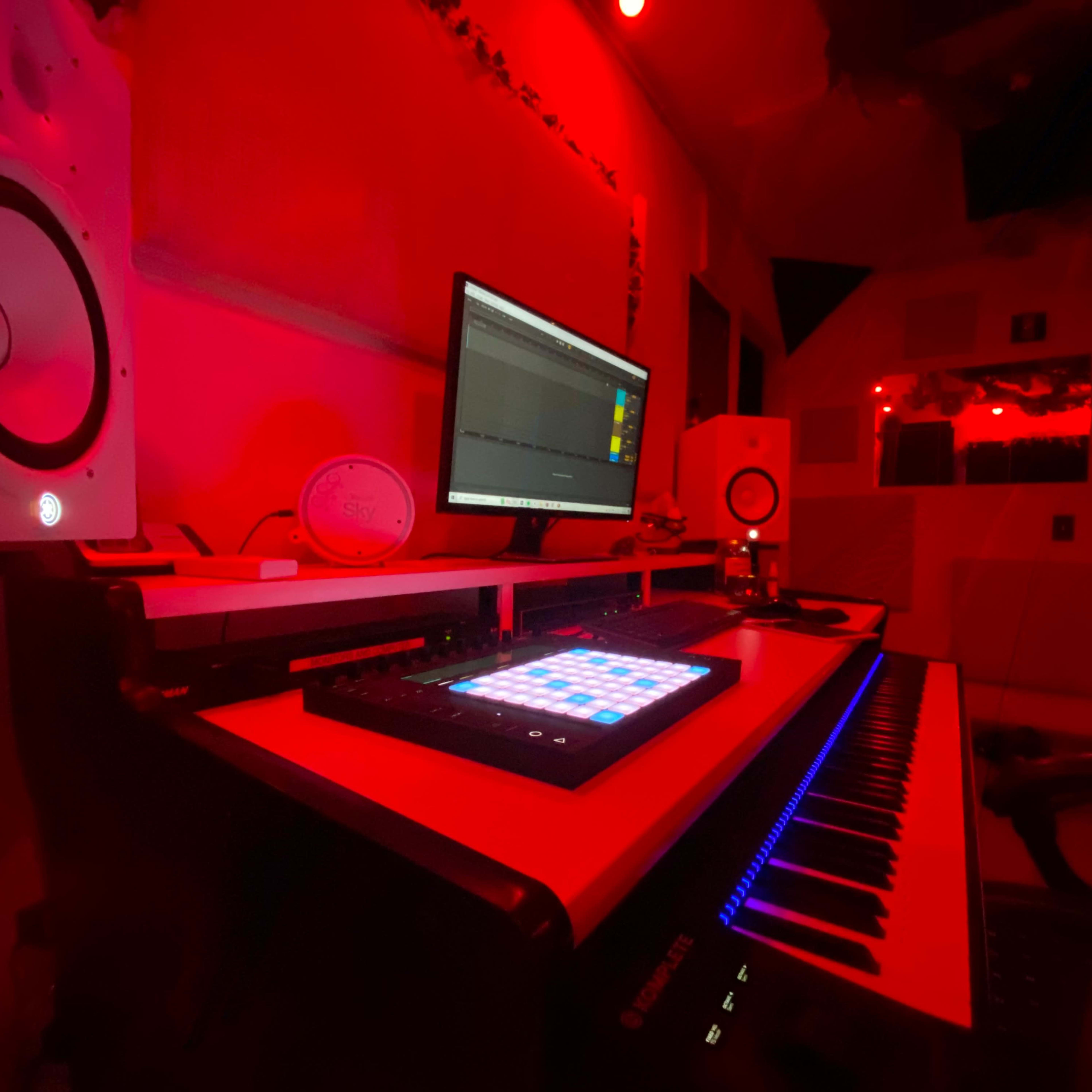 A music studio setup with a computer, a MIDI keyboard, and studio monitors illuminated by red lighting.