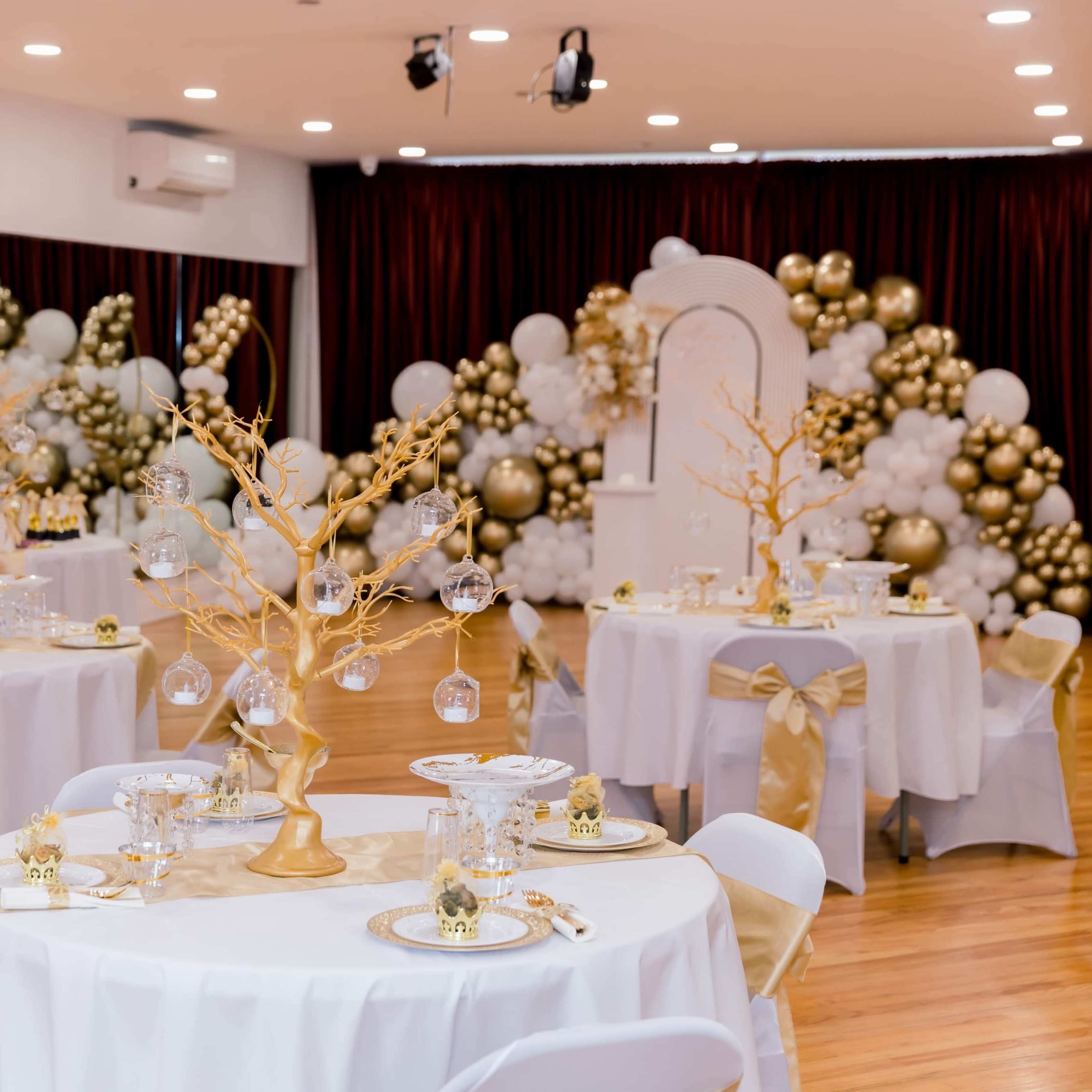An elegantly decorated hall with tables set for an event, featuring white tablecloths and gold accents, surrounded by balloon decor in shades of gold and white.