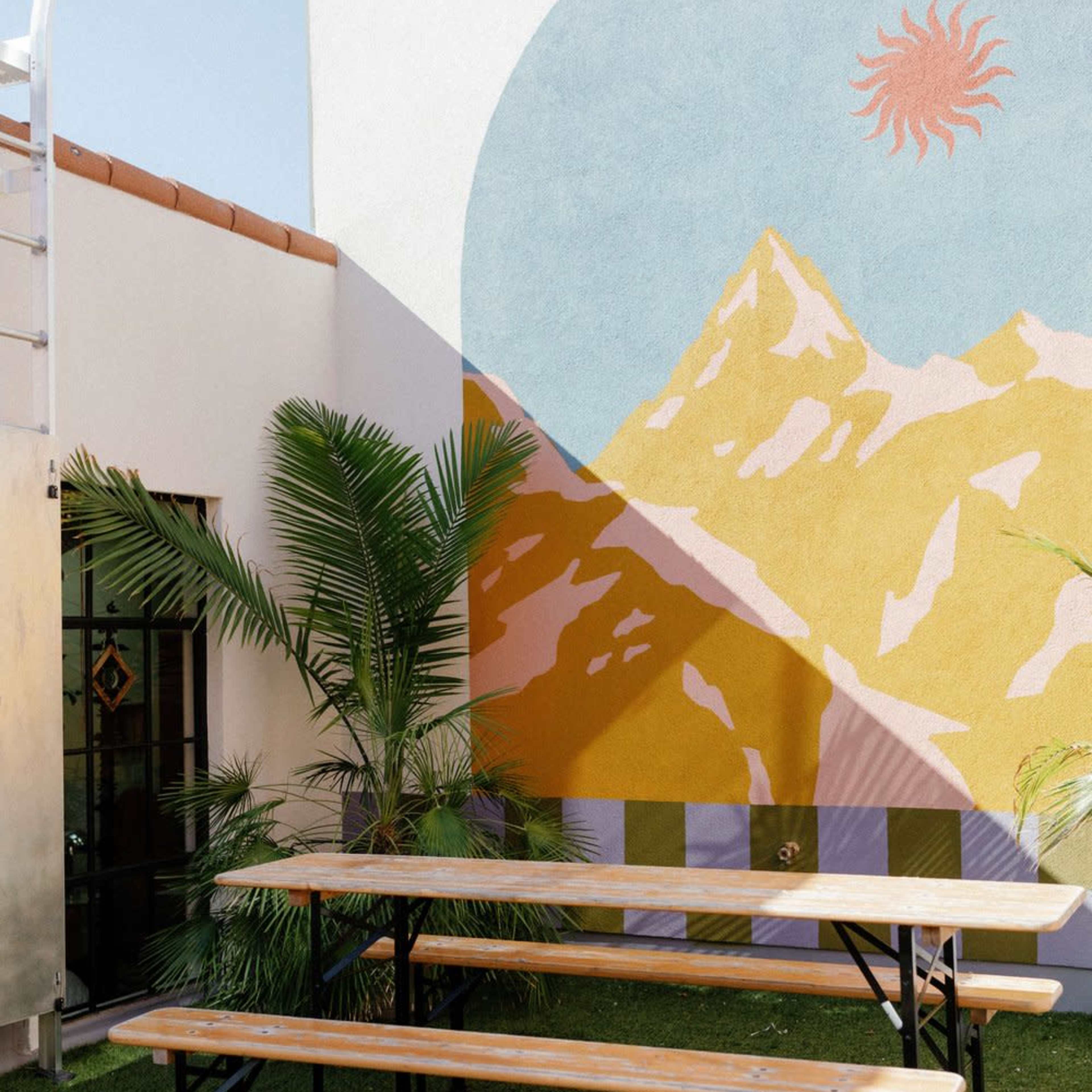 A bright outdoor space features wooden benches in front of a colorful mountain mural, with a lush palm plant nearby.