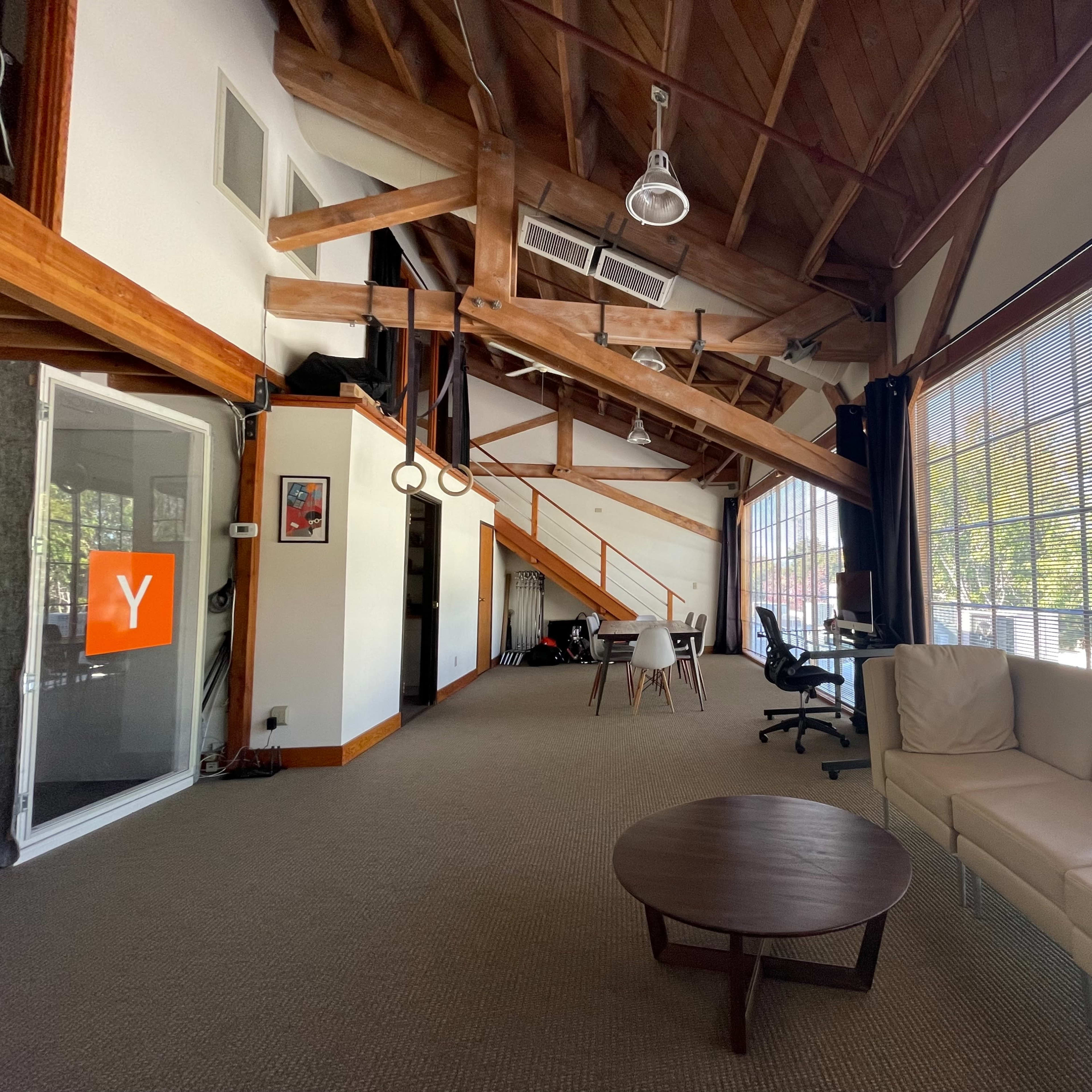 A spacious room with exposed wooden beams, large windows, minimal furniture including a couch and a coffee table, and office workstations in the background.