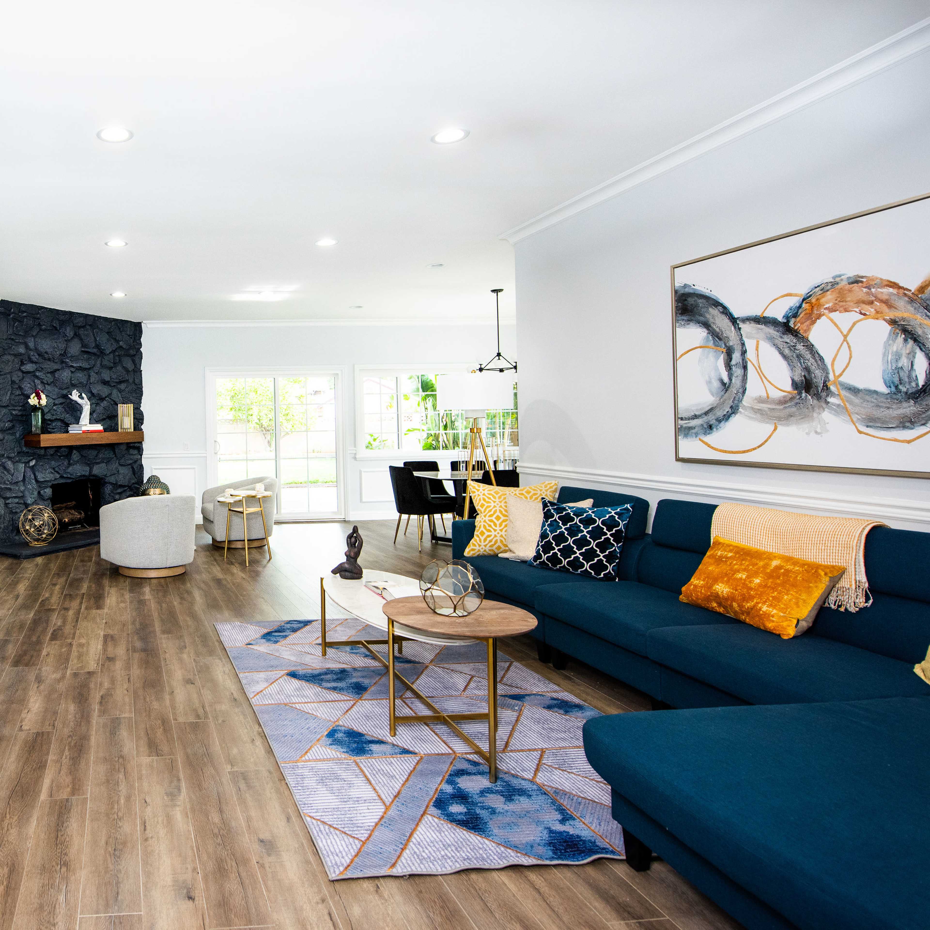 A modern living room featuring a blue sectional sofa, a wooden coffee table, a stone fireplace, and large windows that bring in natural light.