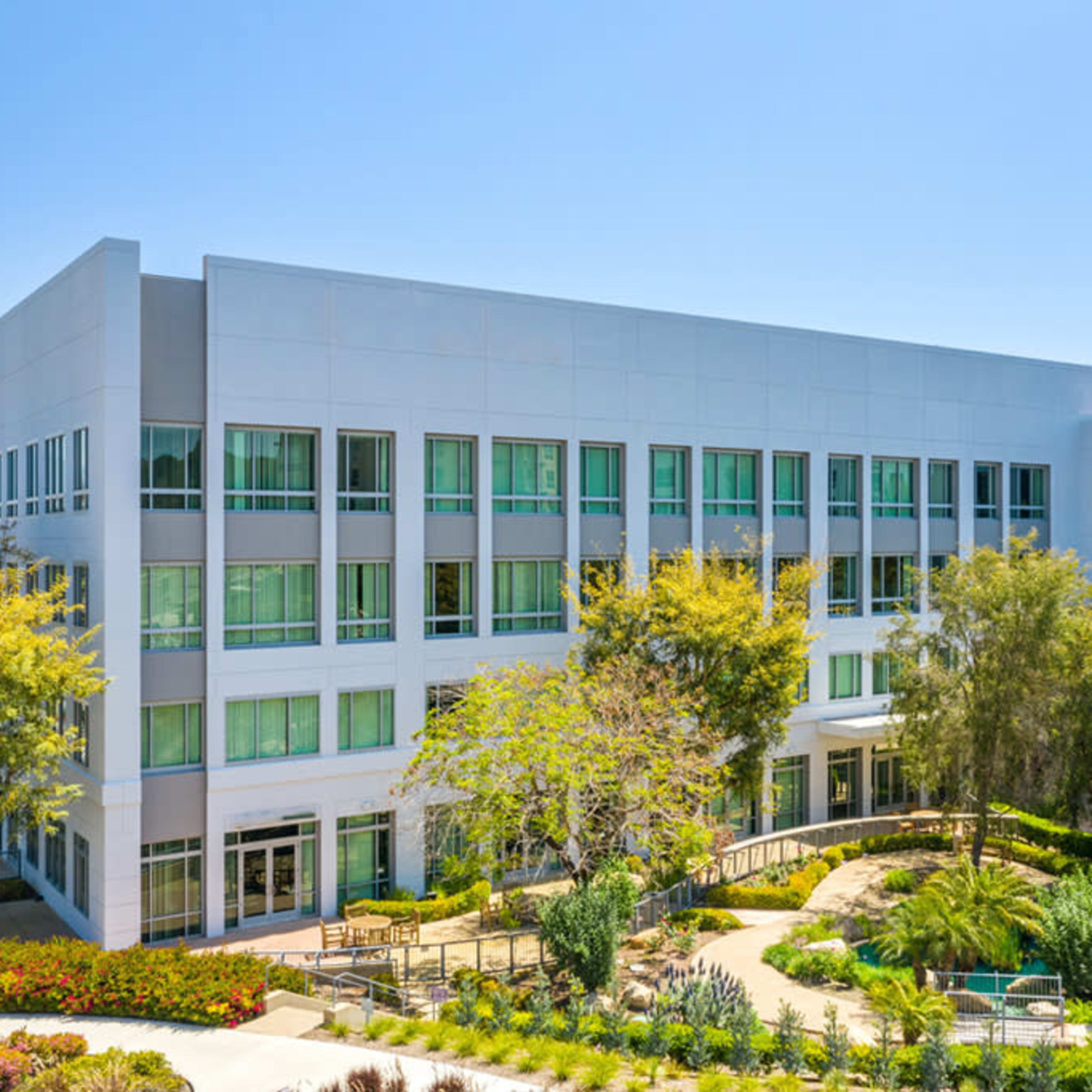 A modern office building with multiple windows is surrounded by landscaped greenery and trees.