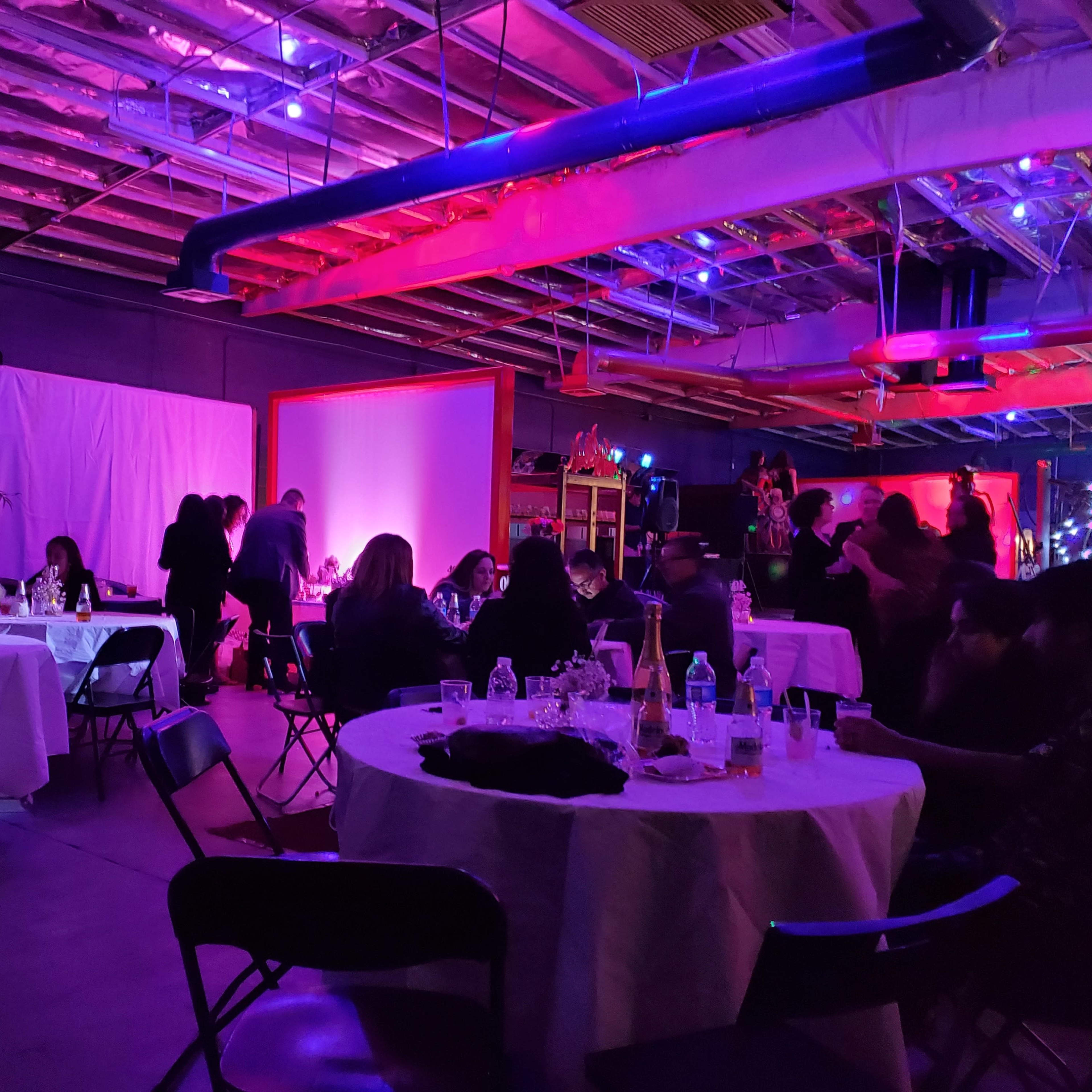 A dimly lit venue features several tables with guests dining, surrounded by colorful lights and a backdrop of plants.