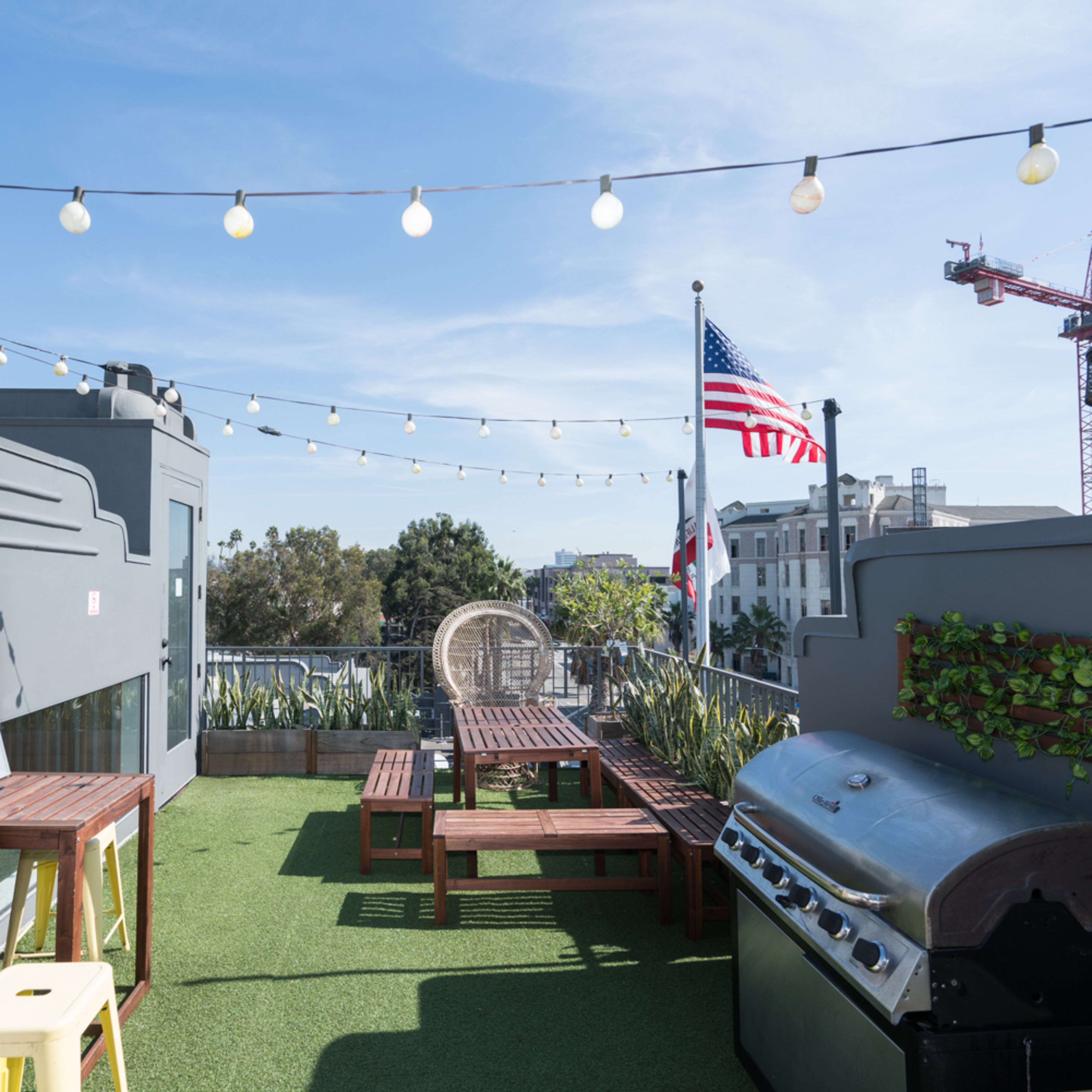 A rooftop area features string lights, seating arrangements, a barbecue grill, and a view of a flag and nearby buildings.