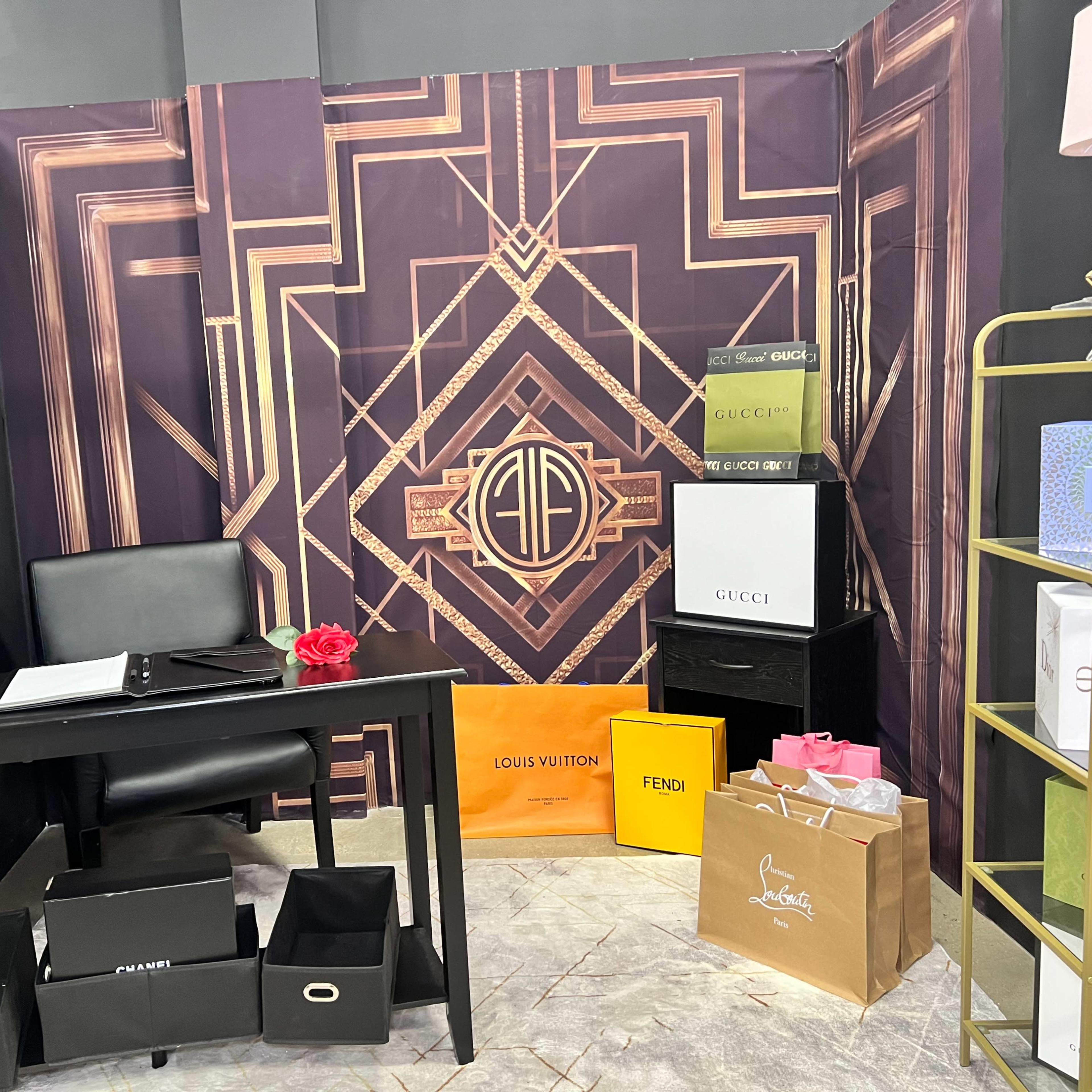 A small room with a desk, a black chair, and various designer brand shopping bags arranged against a decorative backdrop featuring geometric patterns.