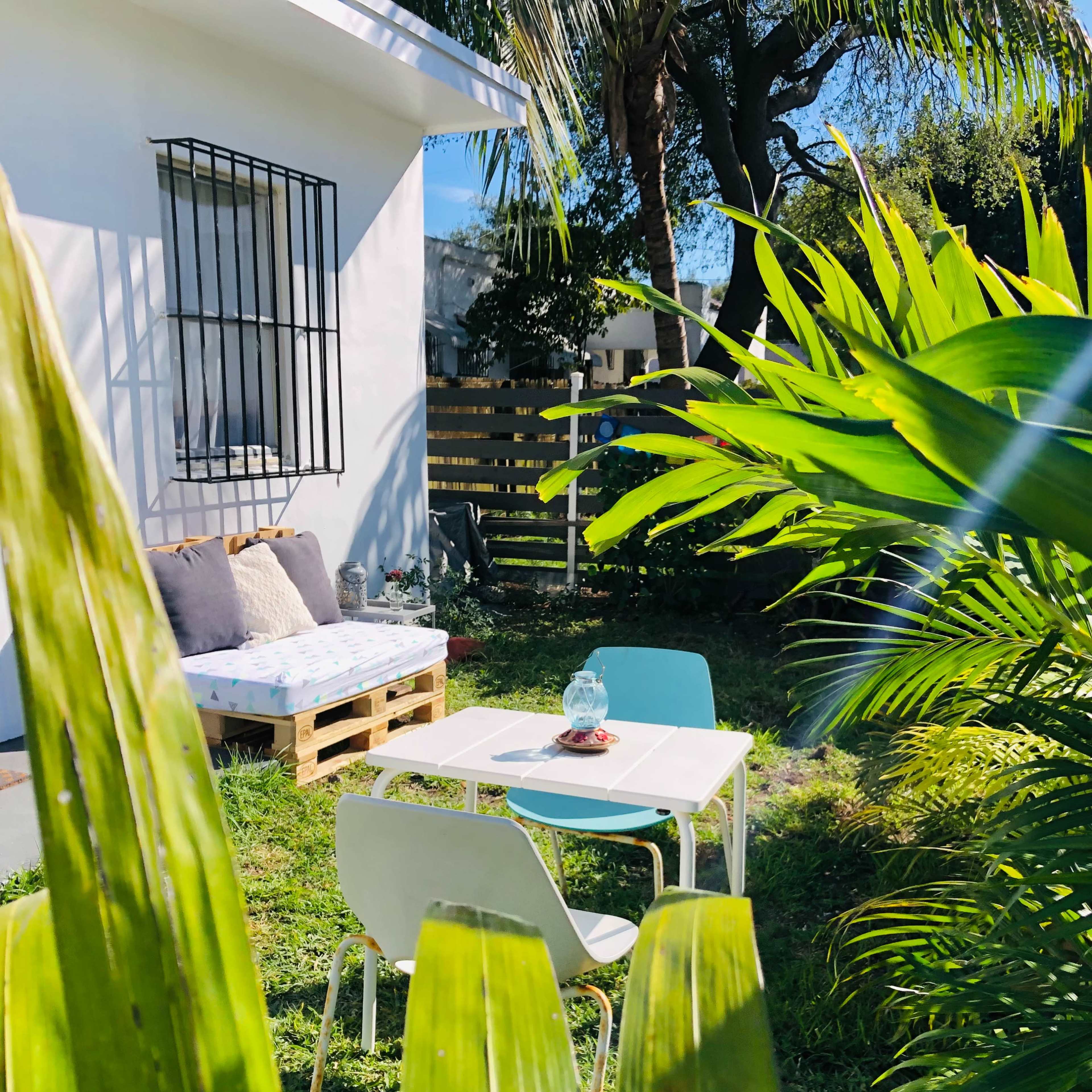 A small outdoor seating area is set up with a table and two chairs, surrounded by lush greenery and a white building in the background.