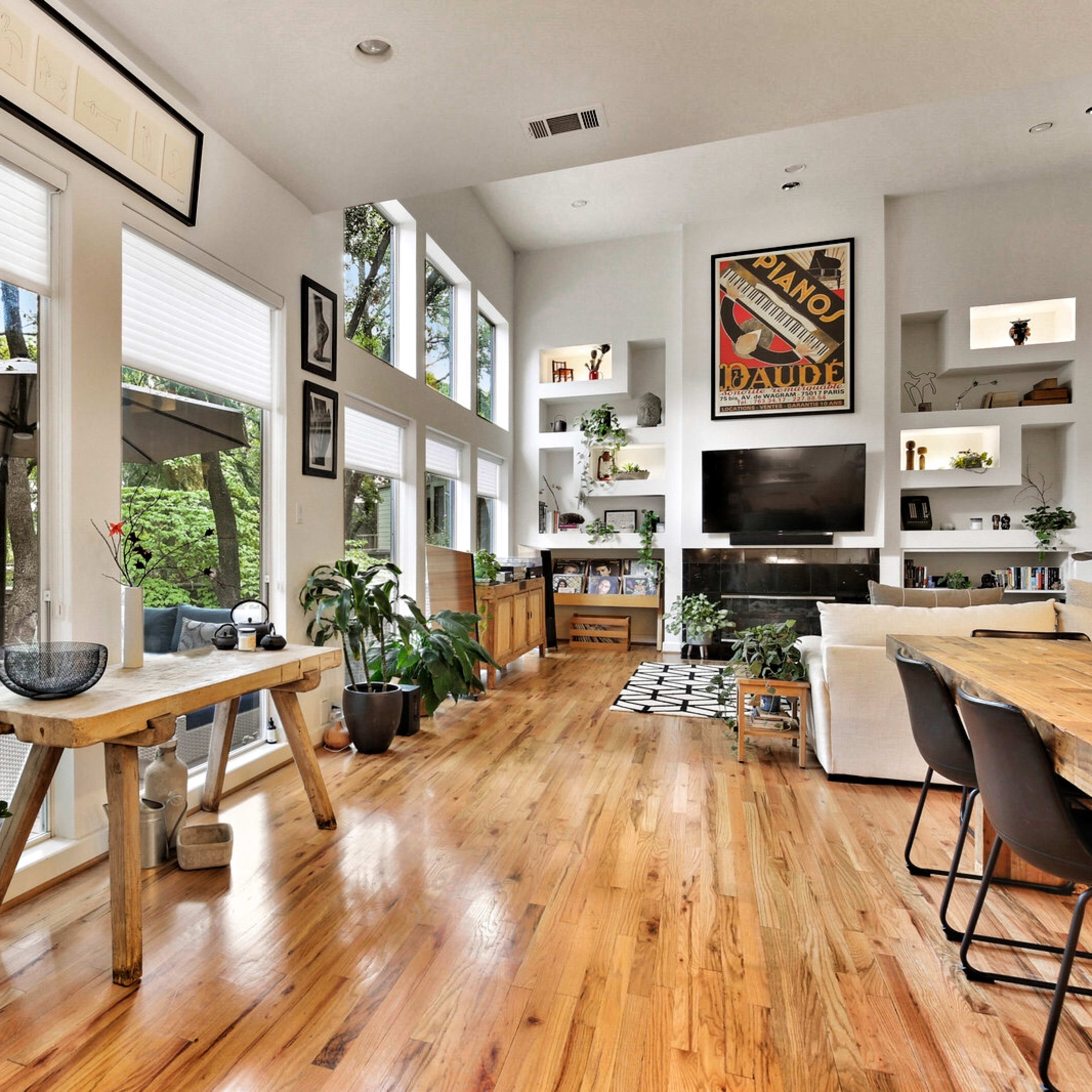The room features large windows, wooden flooring, and a mix of furniture, including a dining table and a seating area with plants and decorative items.