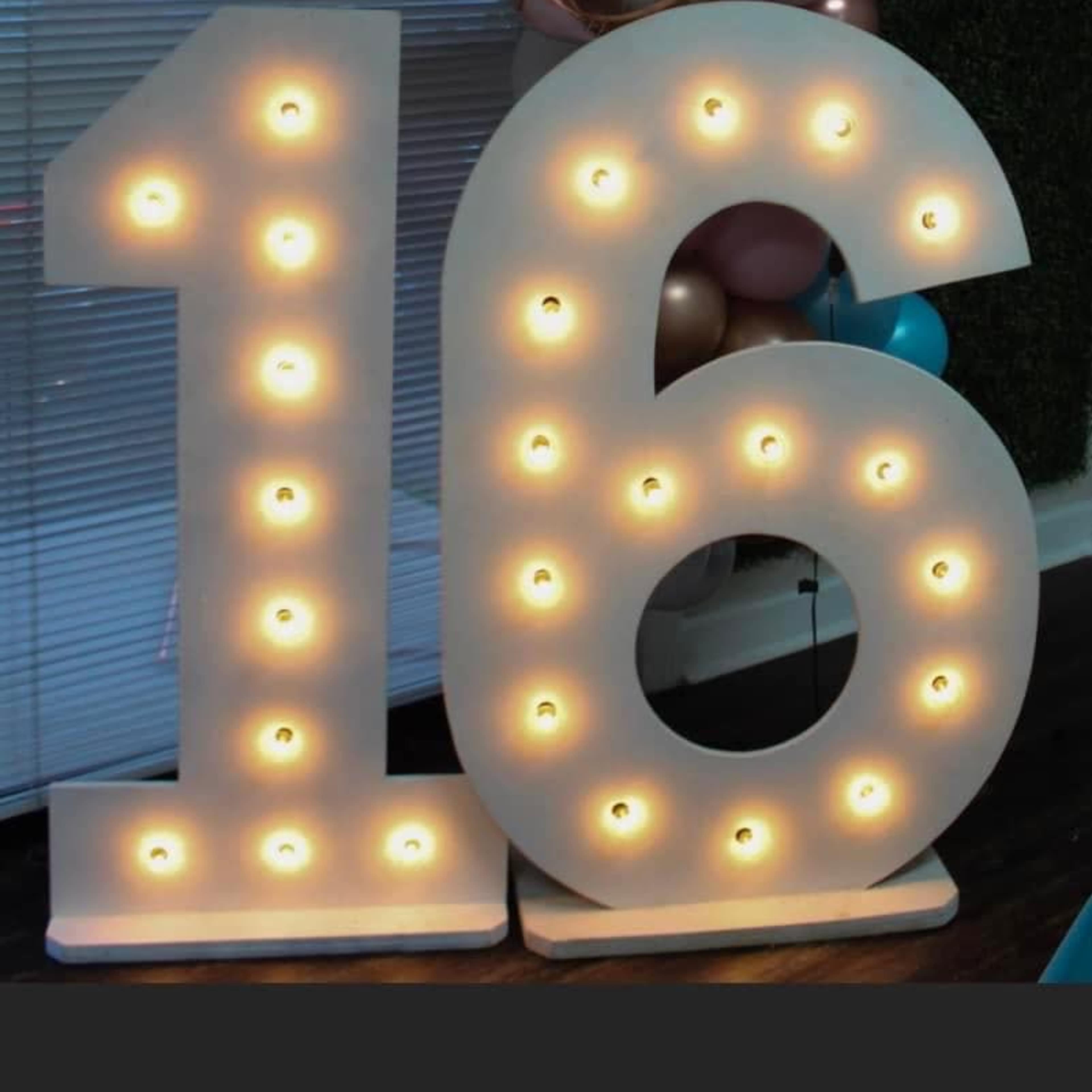 Large illuminated numbers "16" made of white material with bulbs, set against a backdrop of balloons and a wall.