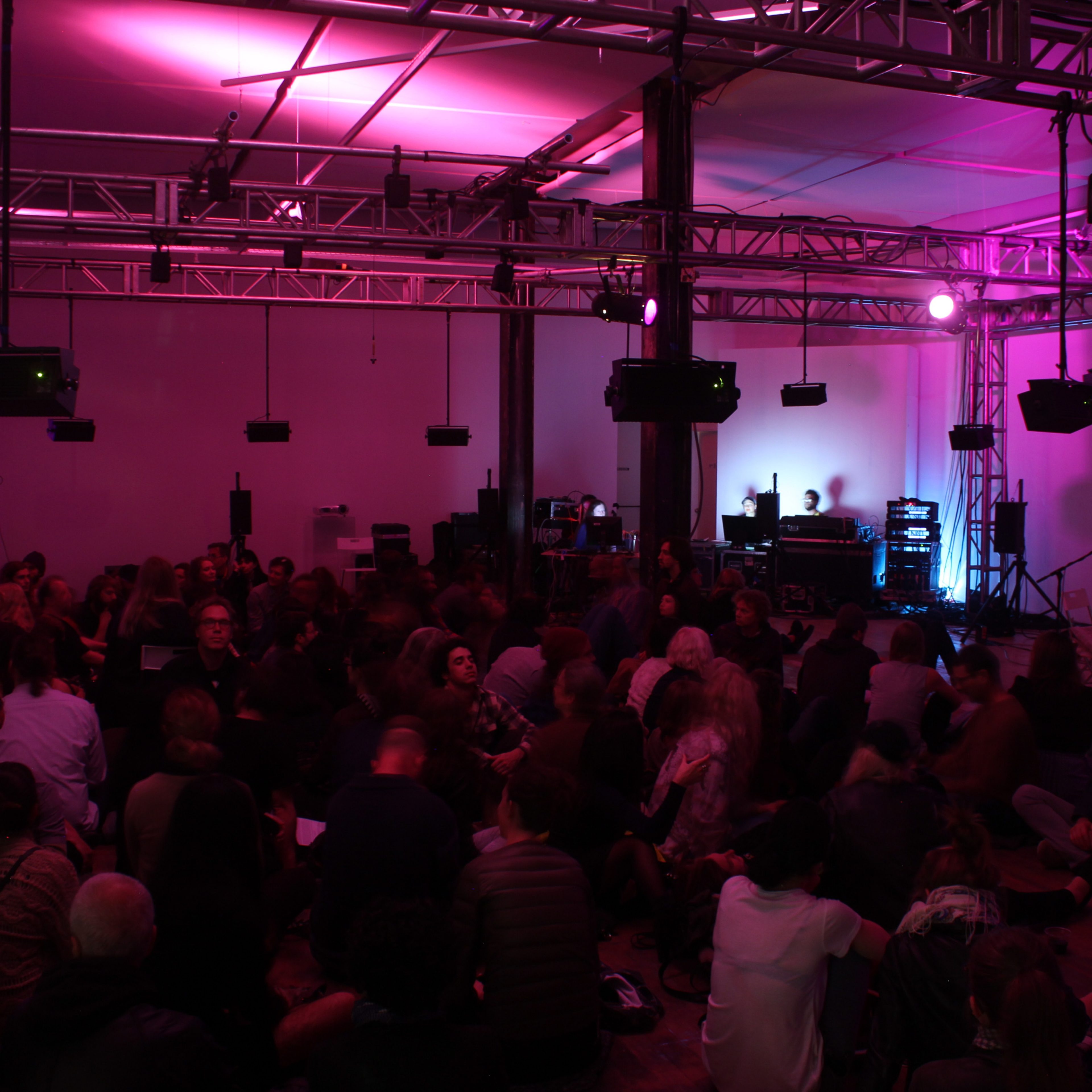 A crowd is seated on the floor of a dimly lit venue with purple lighting and musical equipment set up on a stage.