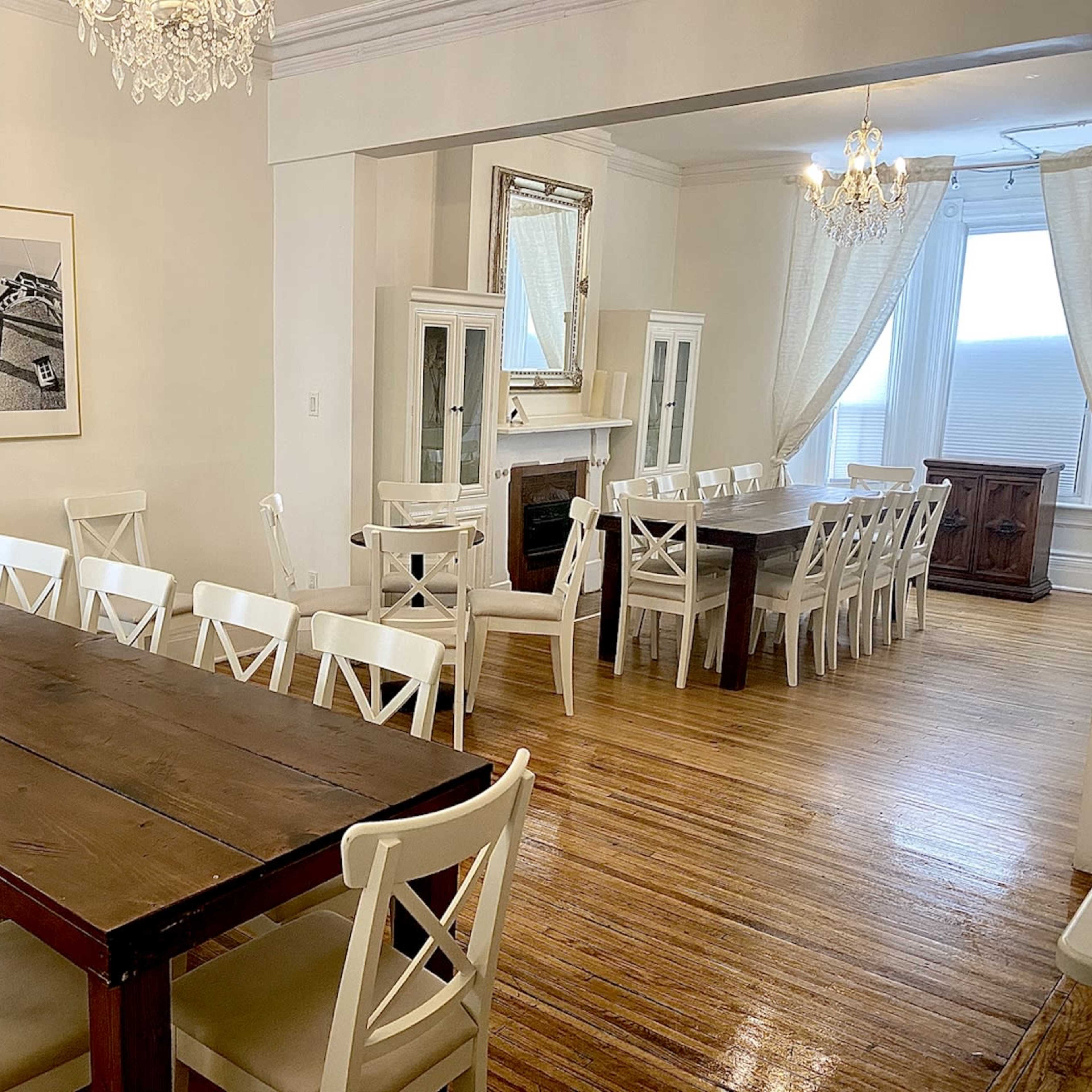 A spacious dining area with two long wooden tables surrounded by white chairs, complemented by large windows and light-colored walls.