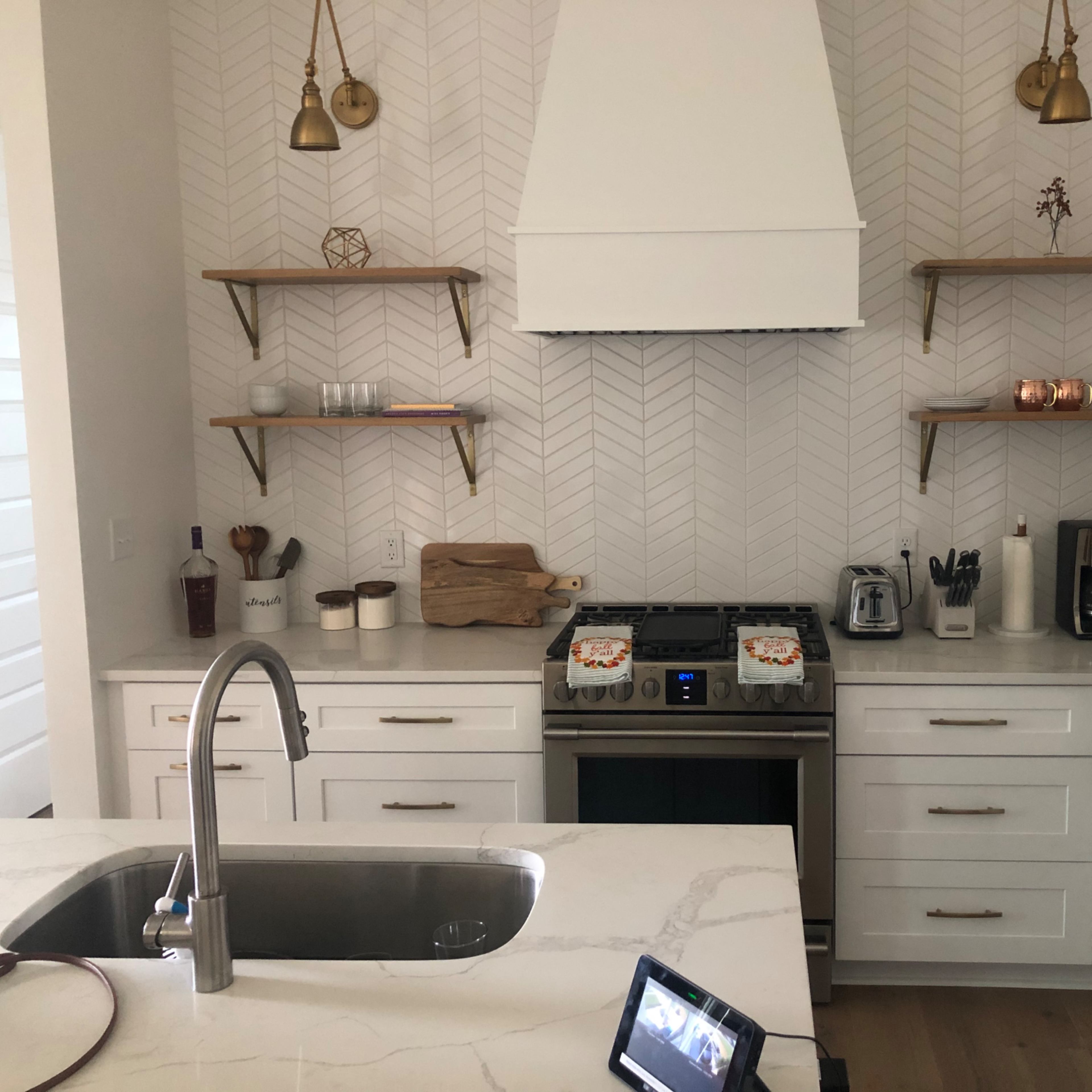 A modern kitchen features a central island with a sink, stainless steel appliances, and herringbone-patterned wallpaper, along with open shelving displaying various kitchen items.