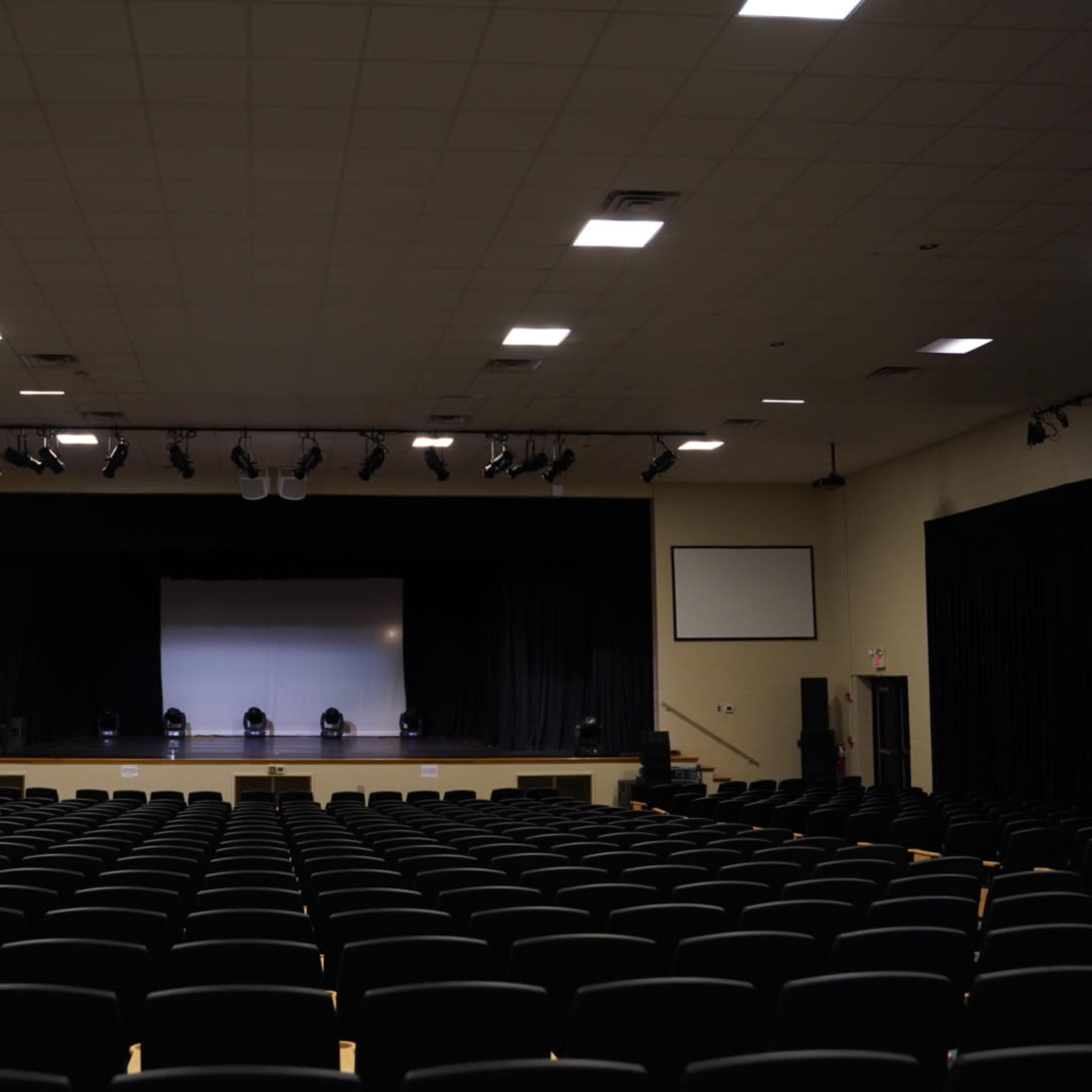 A large auditorium with multiple rows of black seats facing a stage that has a central screen and spotlights.