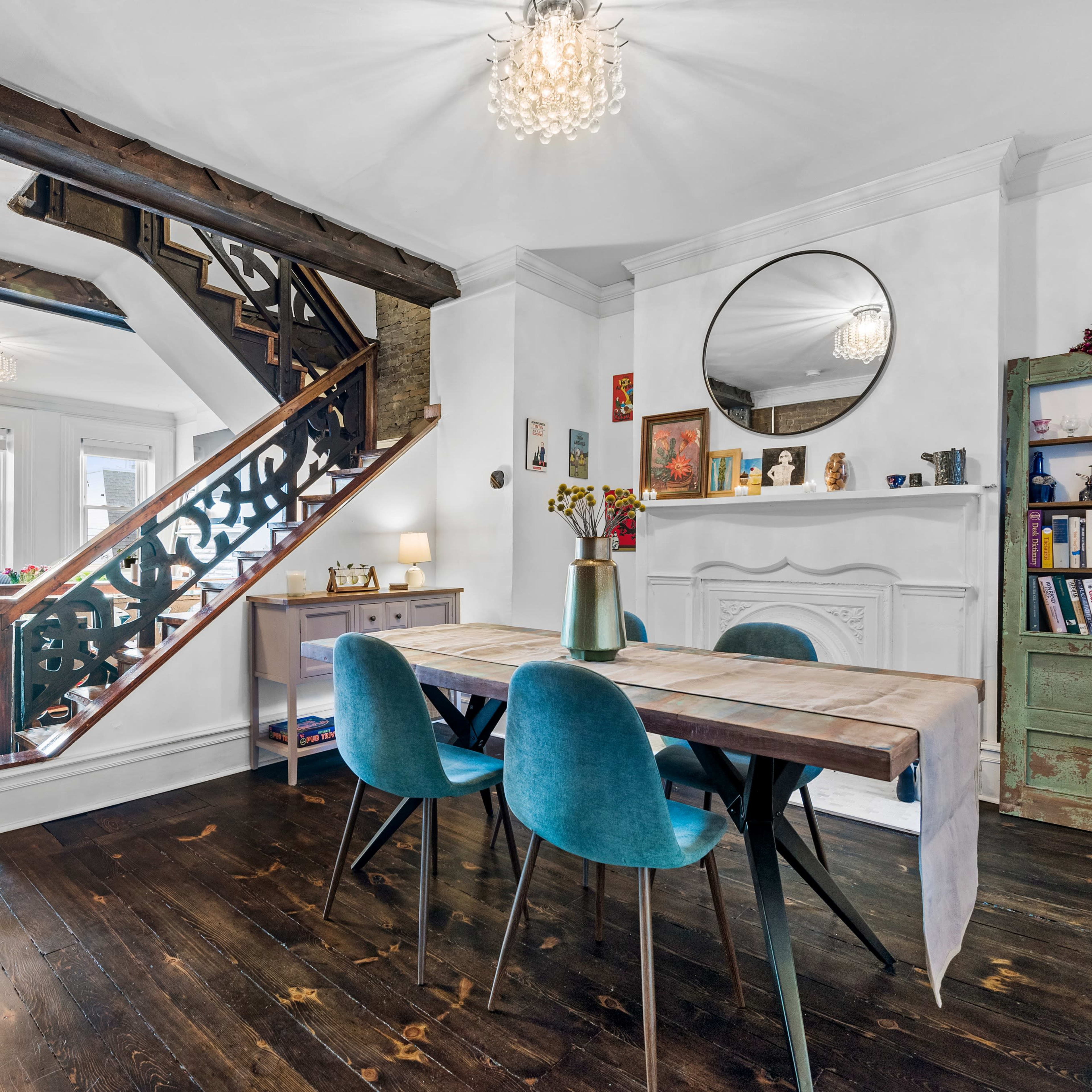 A well-decorated interior space featuring a wooden dining table, blue chairs, and a staircase with decorative railing, accompanied by various pieces of furniture and decor against a backdrop of large windows.