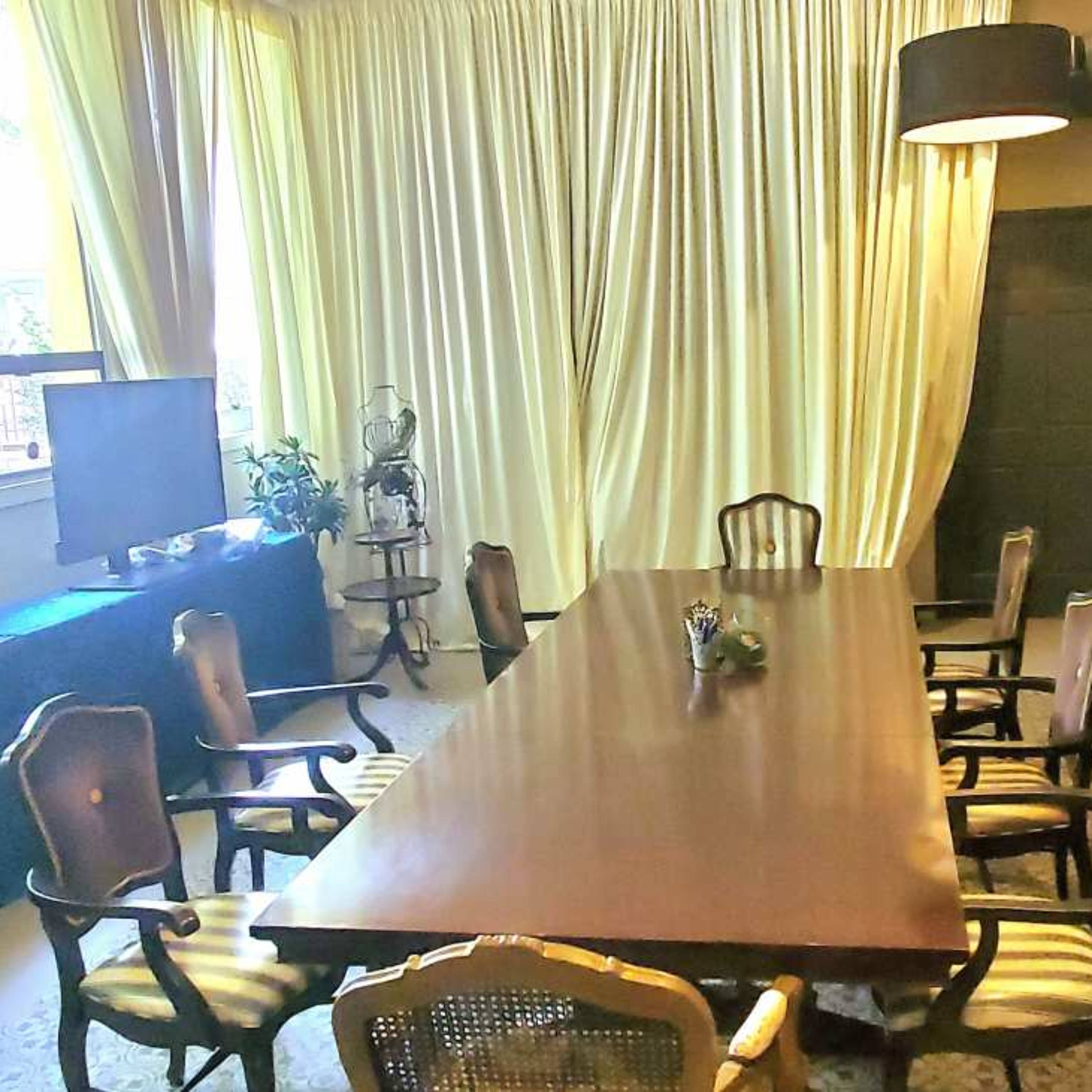 A conference room is arranged with a long wooden table surrounded by eight chairs, with curtains and two television screens in the background.