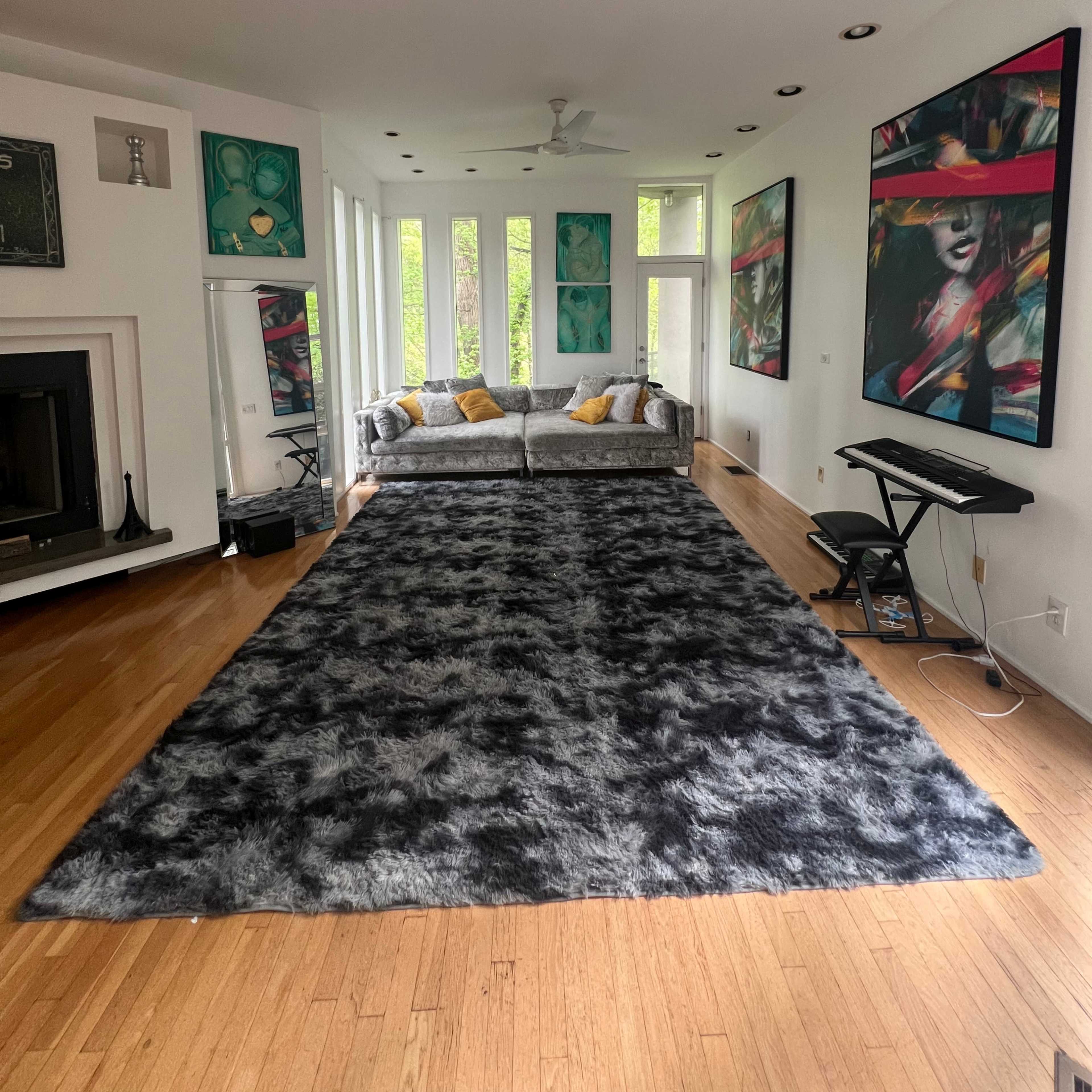 A spacious living room featuring a gray sofa with yellow cushions, a large black and gray area rug, and tall windows with green foliage outside.