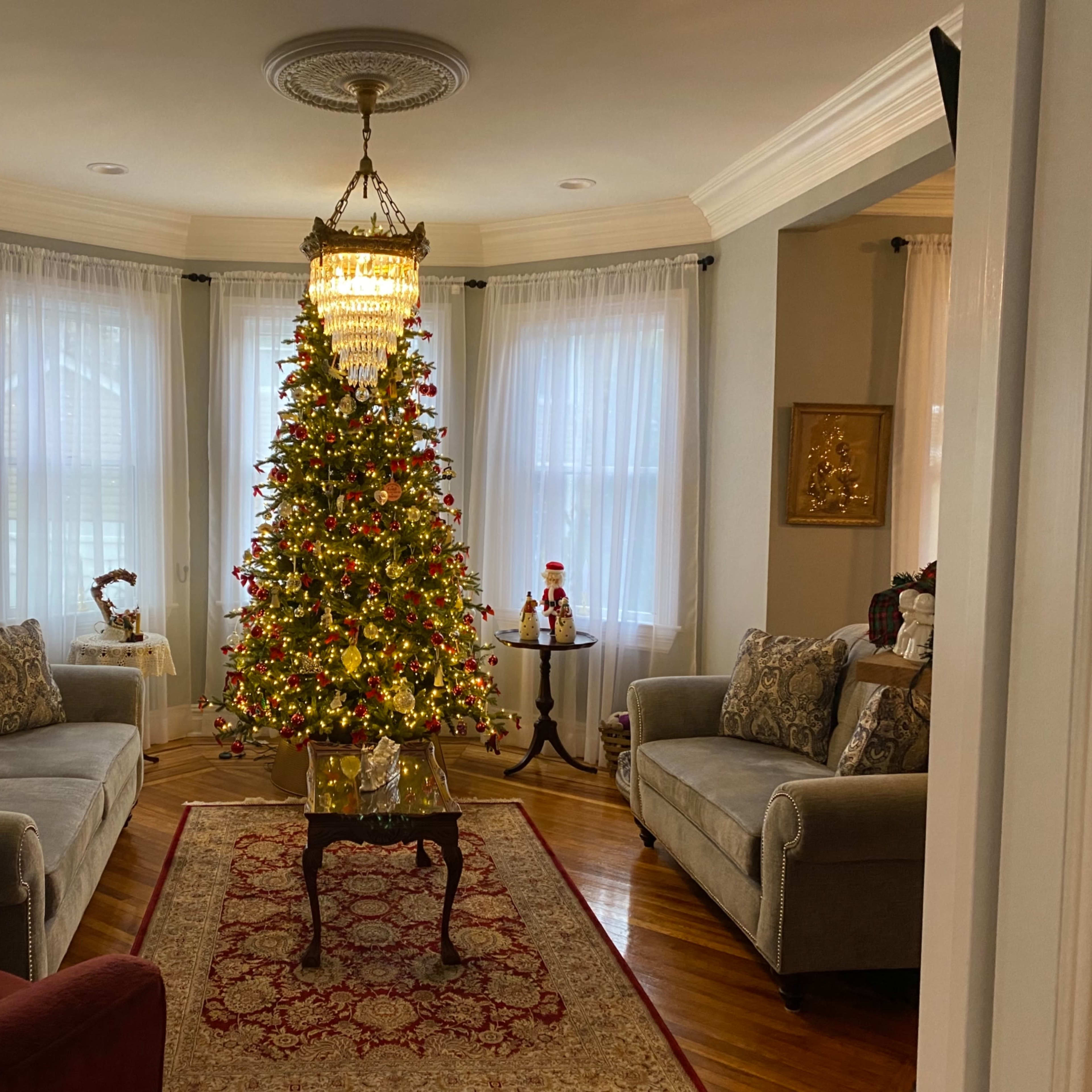A living room features a decorated Christmas tree in the center, flanked by two sofas and a small table, with large windows allowing natural light to enter.