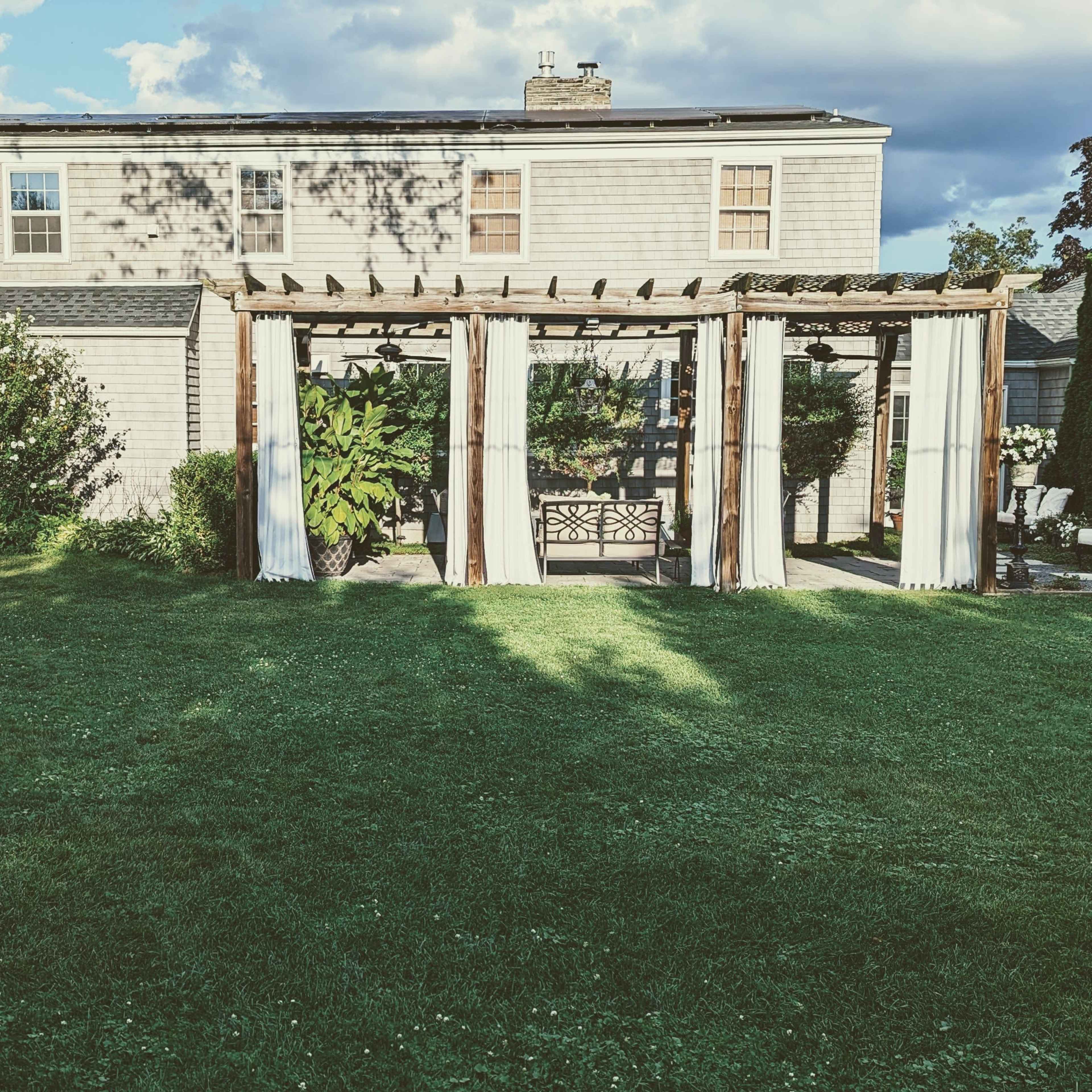 A backyard with a pergola covered in light drapes, grass, and a house in the background under a partly cloudy sky.
