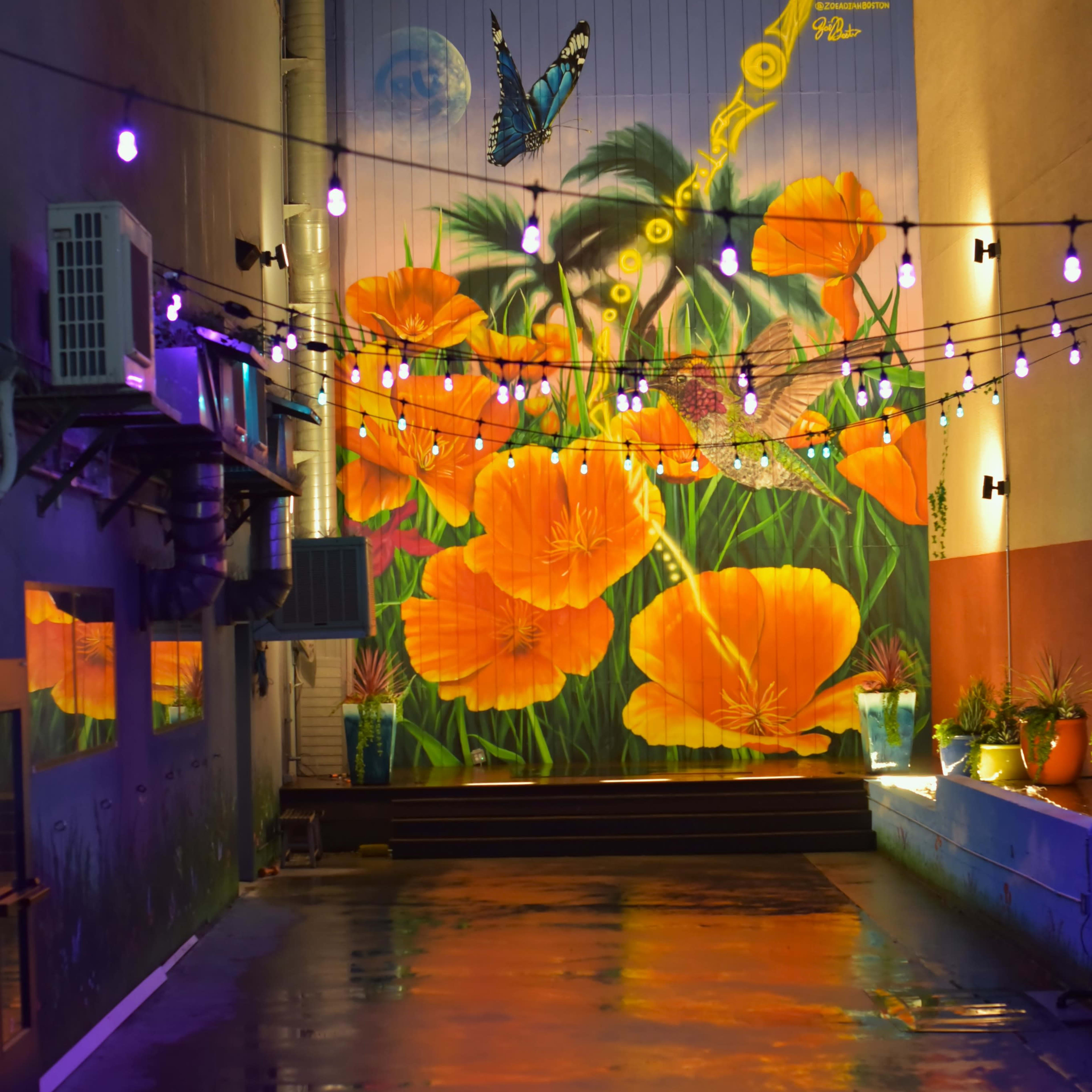 A brightly colored mural of orange flowers and a butterfly on a wall beside a water feature, illuminated by string lights at night.