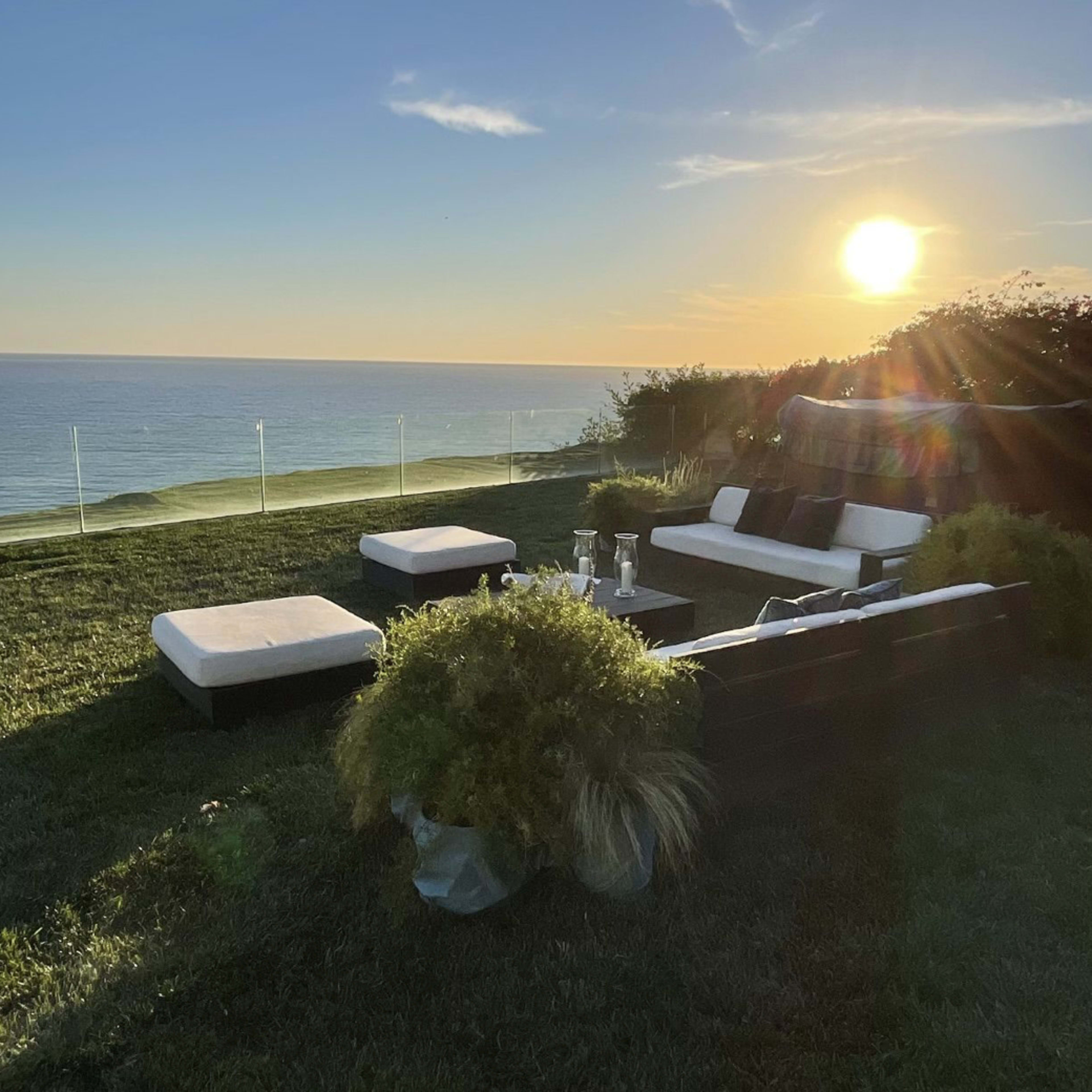 A cozy outdoor seating area overlooks the ocean during sunset, with a clear view of the horizon.