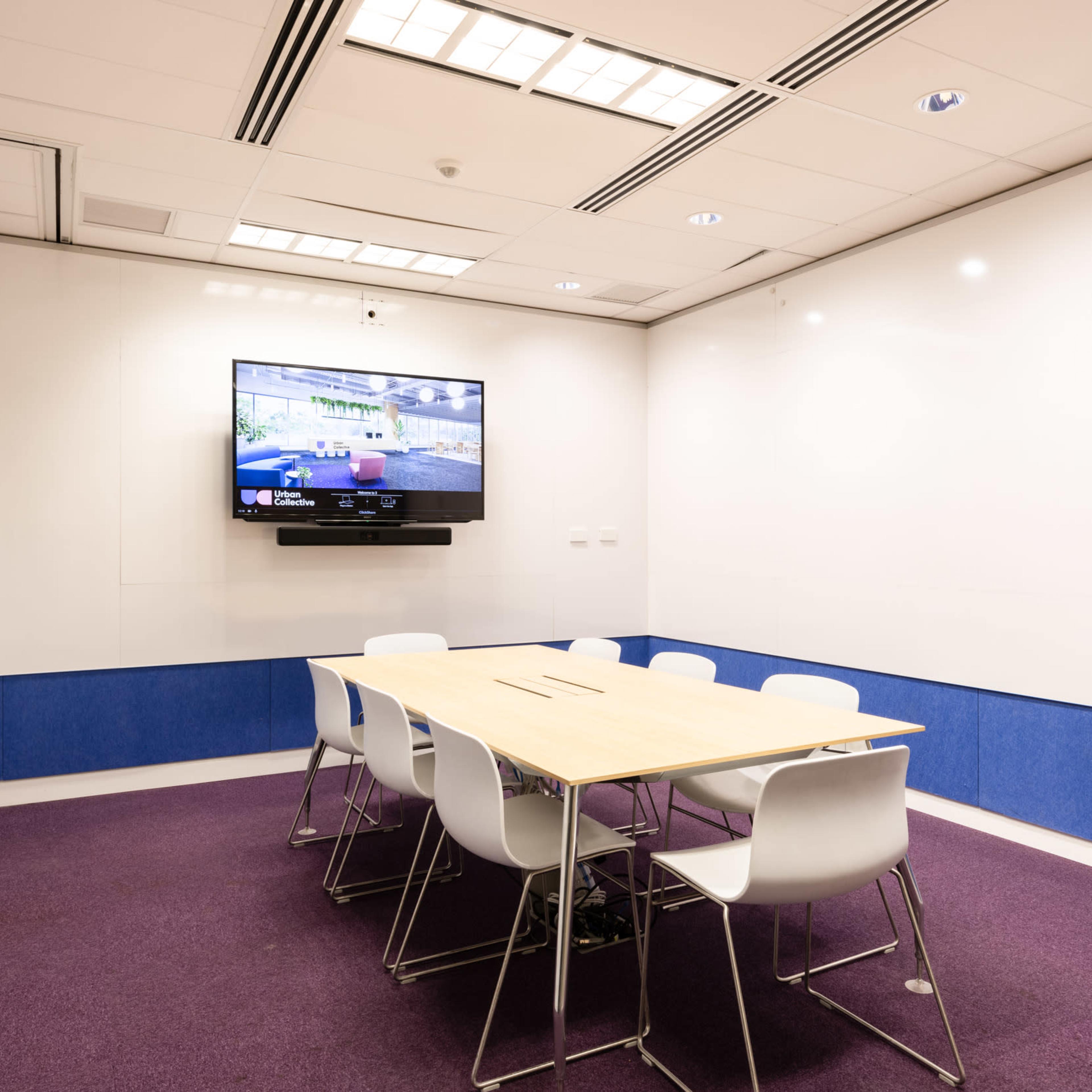 A modern conference room with a wooden table surrounded by white chairs and a wall-mounted television.