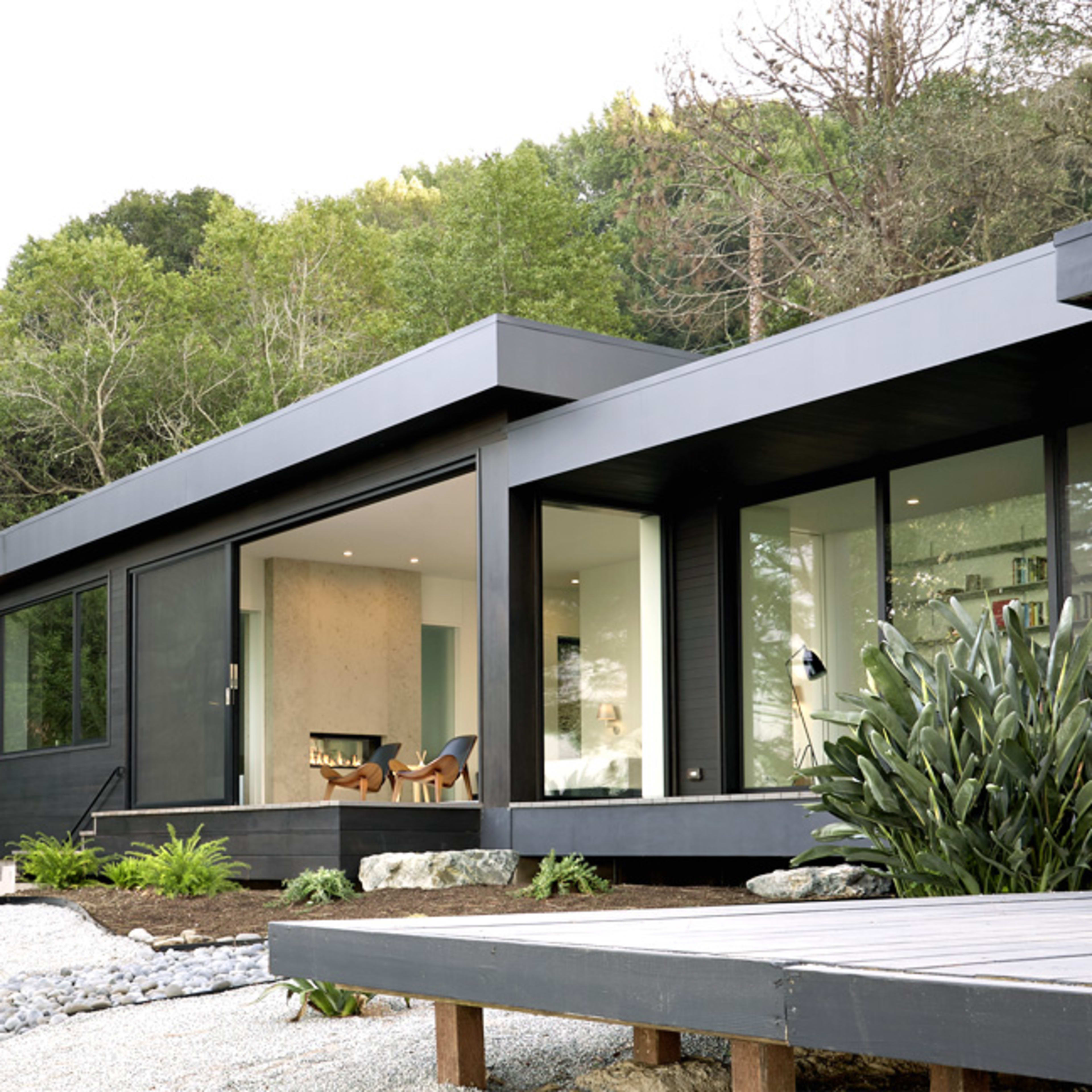 A modern house with large windows and a contrast of black siding and yellow accents overlooks a landscaped yard and surrounding trees.