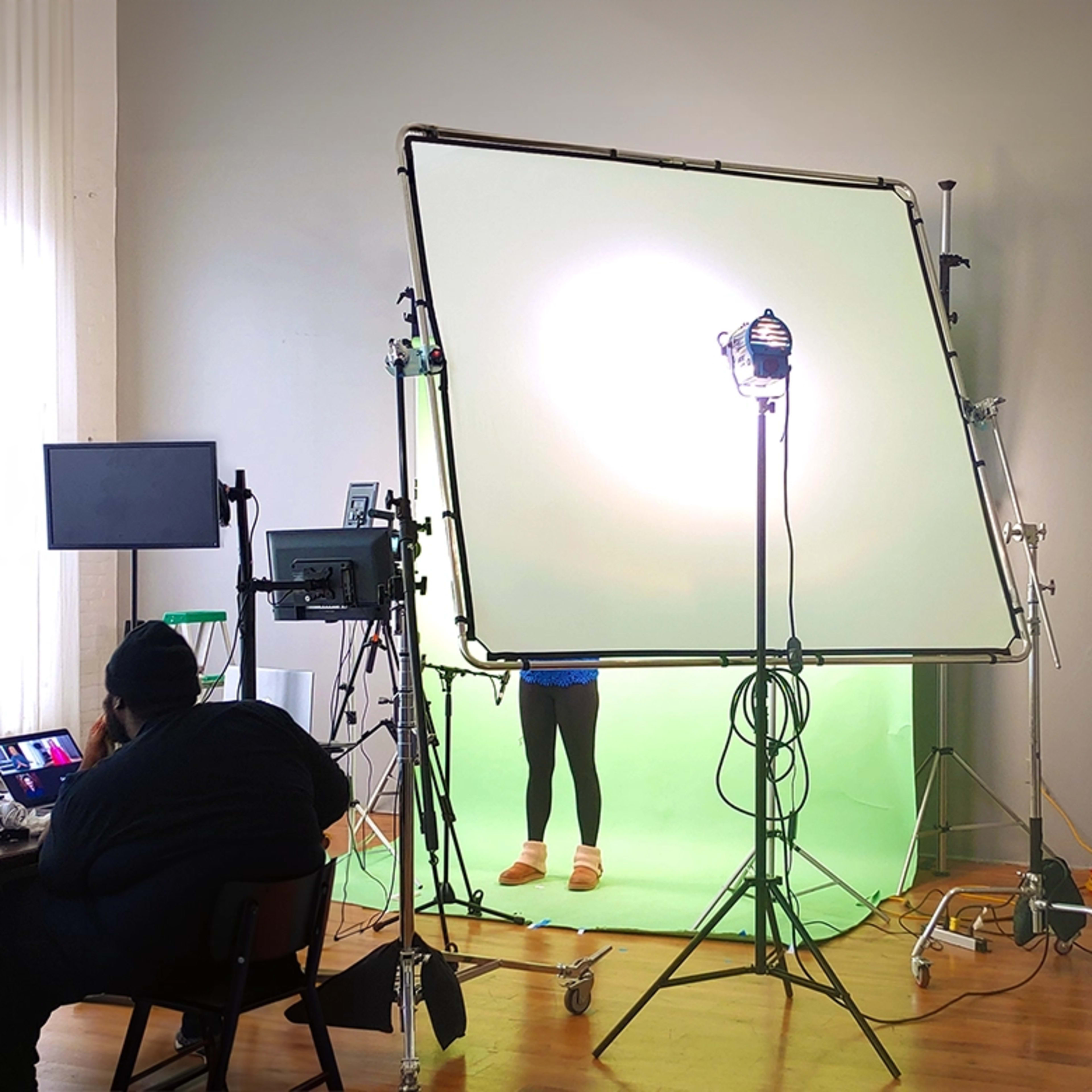 A person sits at a table in a studio, working on a computer in front of a green screen setup with lighting equipment behind them.