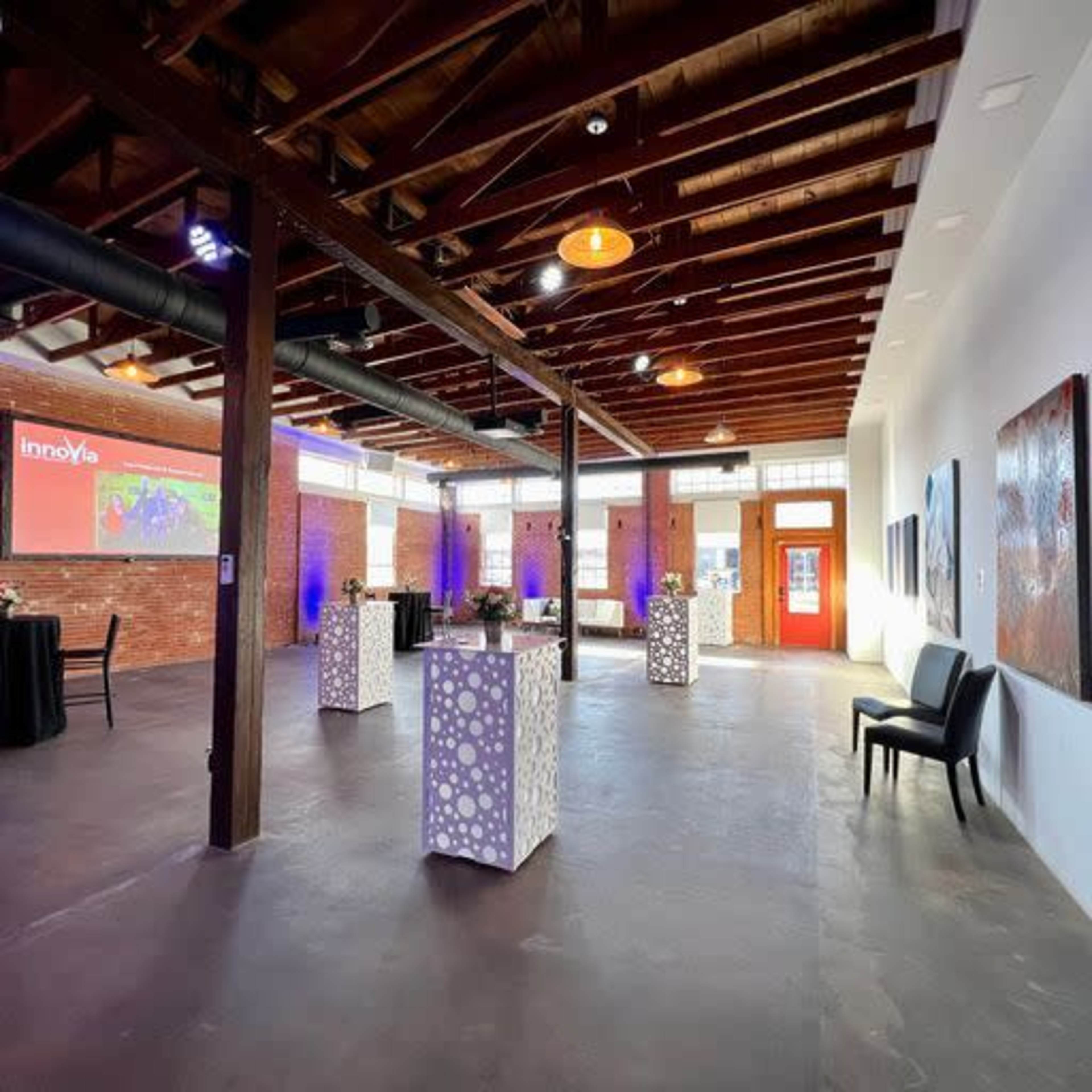 A spacious event venue with high ceilings, wooden beams, and modern decor, featuring several tables with floral arrangements and decorative lighting.
