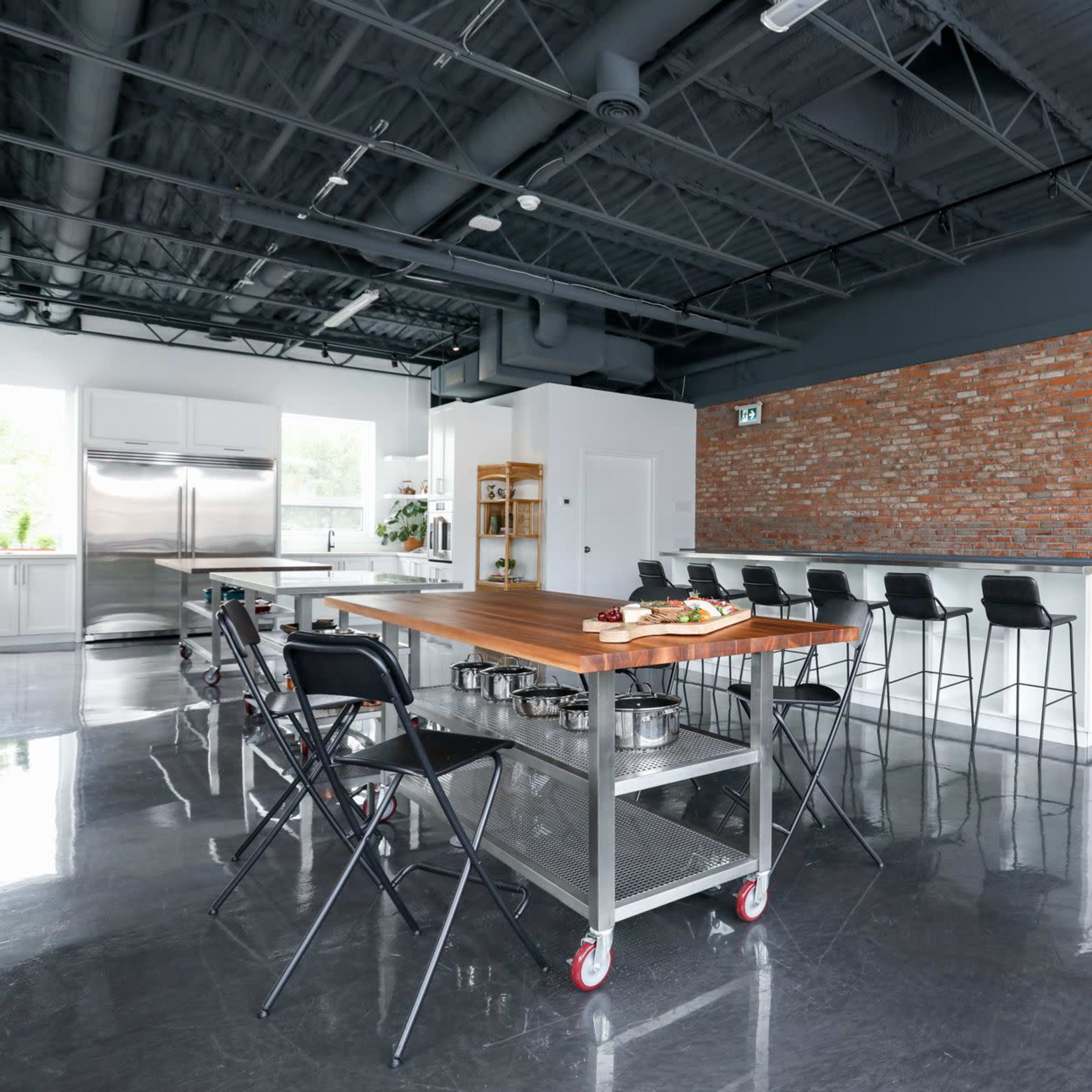 A modern kitchen with a large wooden island, metal stools, and a brick wall, featuring stainless steel appliances and a minimalistic design.