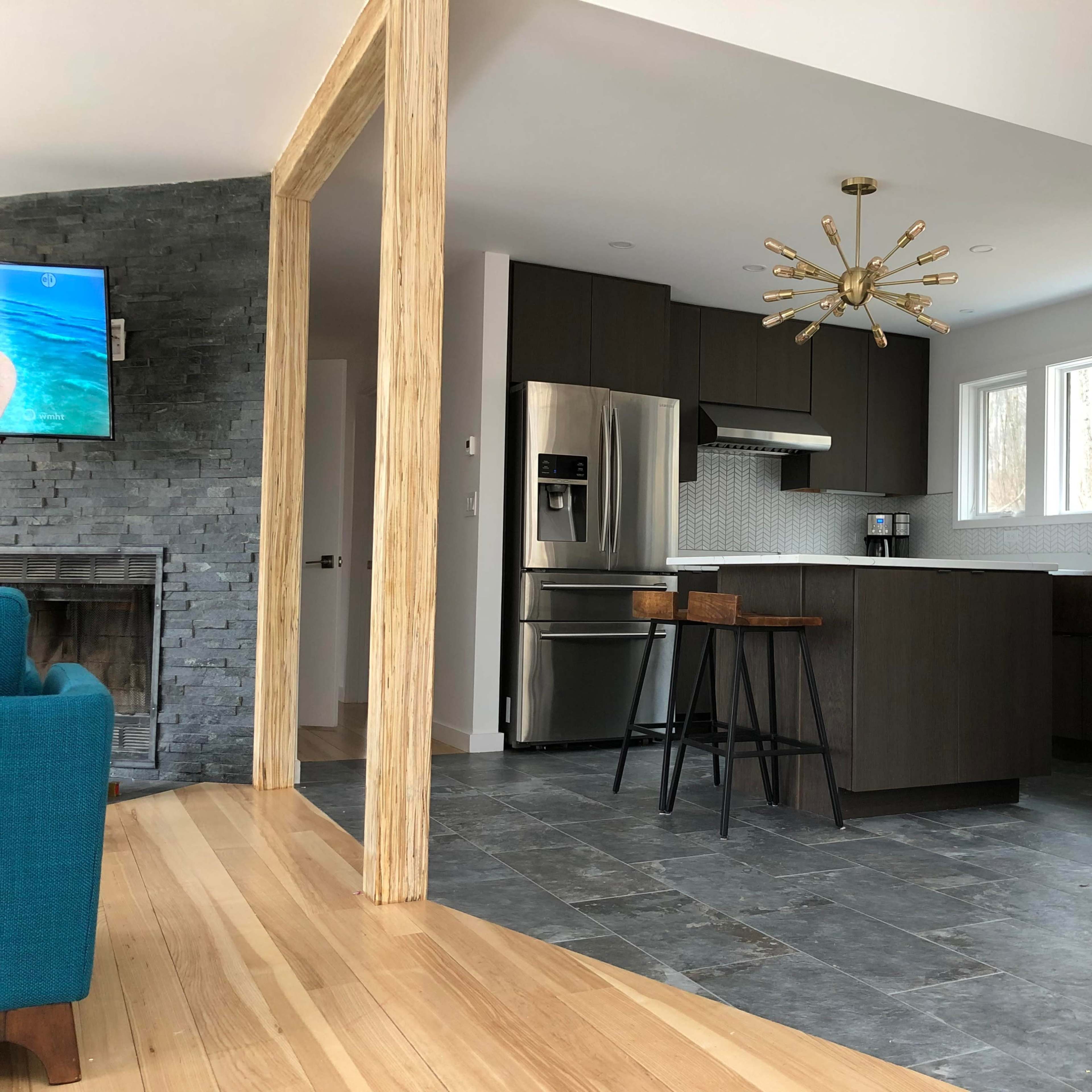 A modern kitchen features dark cabinetry, stainless steel appliances, and large windows, with an adjacent living area visible through a wooden frame.