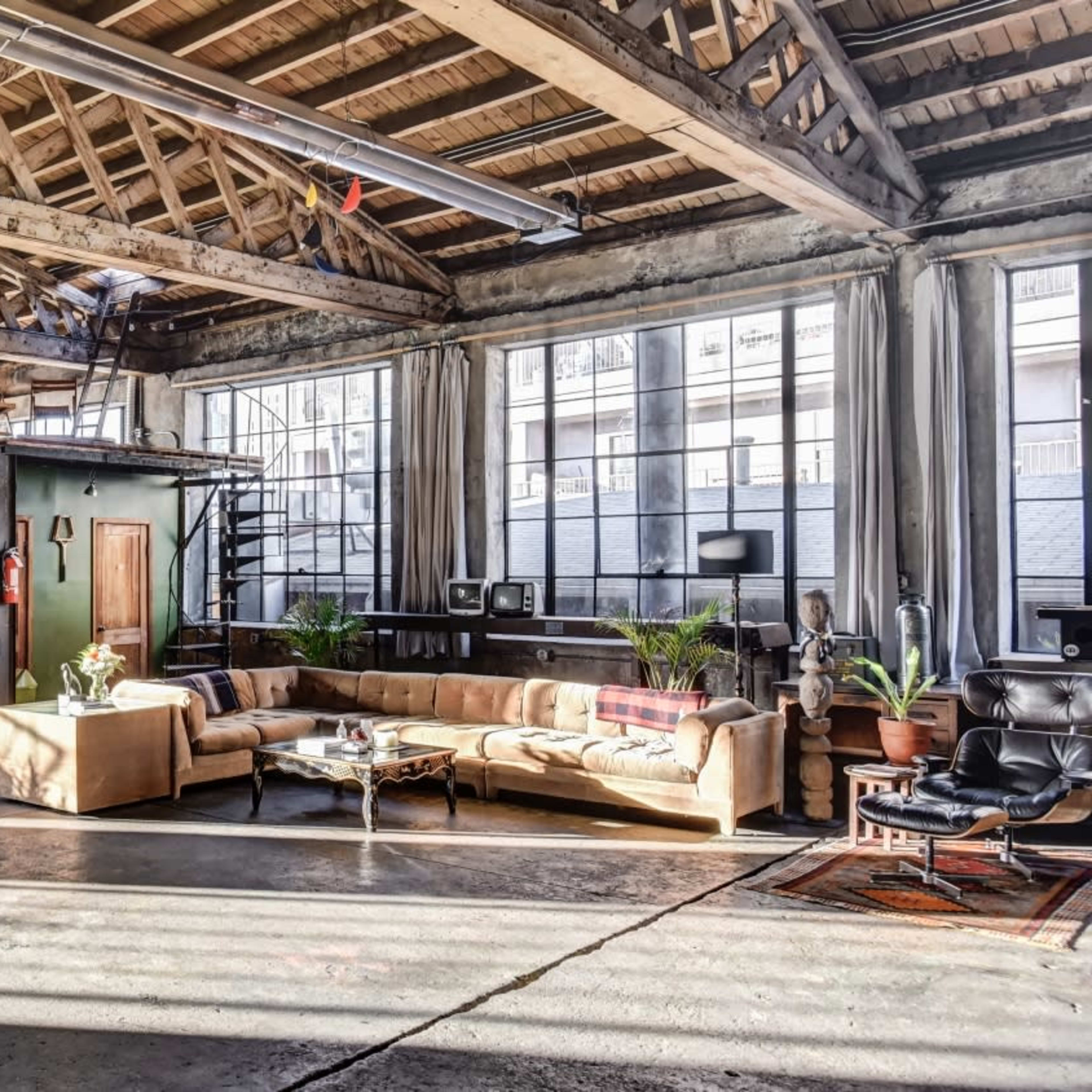 A spacious industrial-style loft with large windows, a seating area, a staircase leading to an upper level, and various plants placed throughout the space.