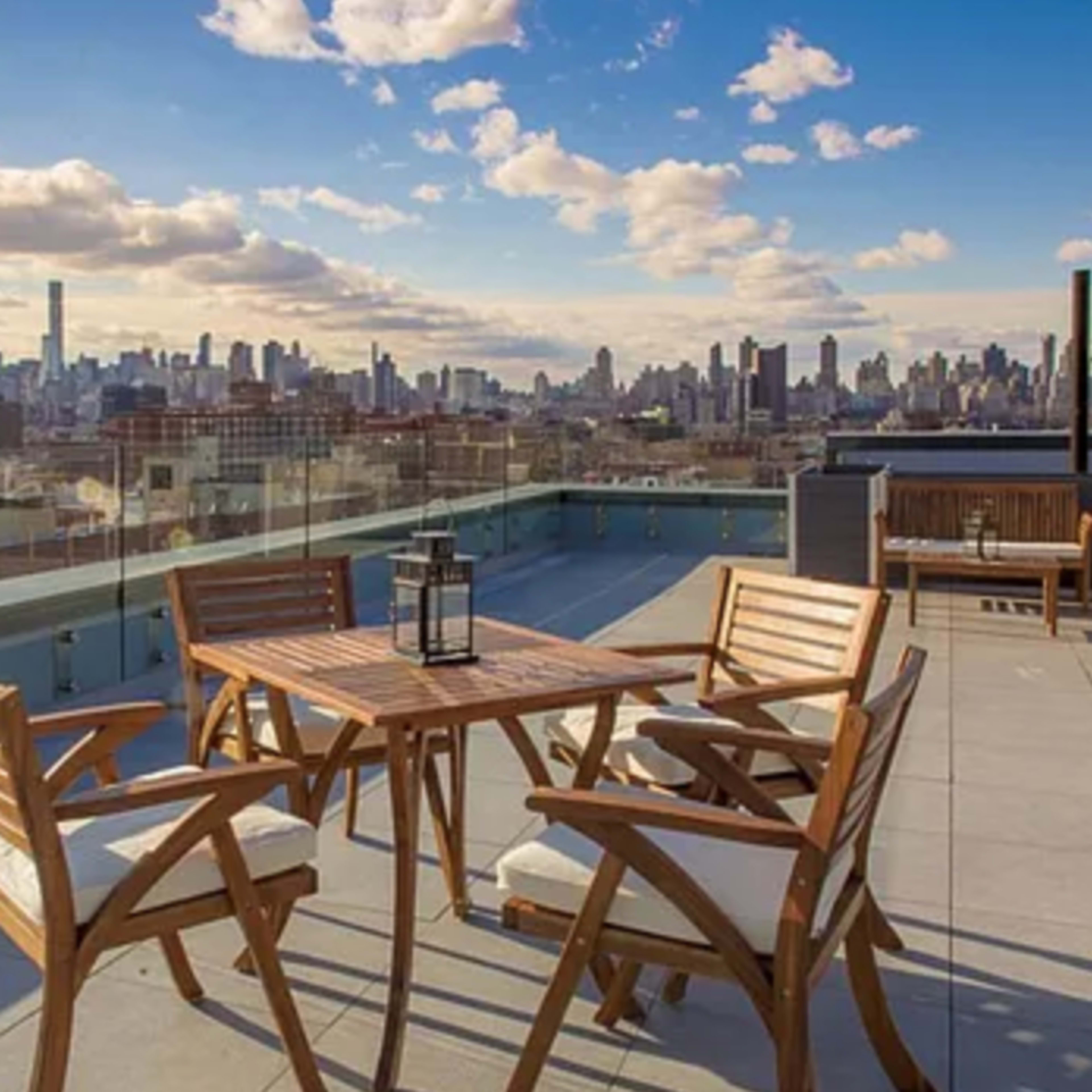 A rooftop terrace features a wooden table surrounded by four chairs, offering a panoramic view of a city skyline under a cloudy sky.