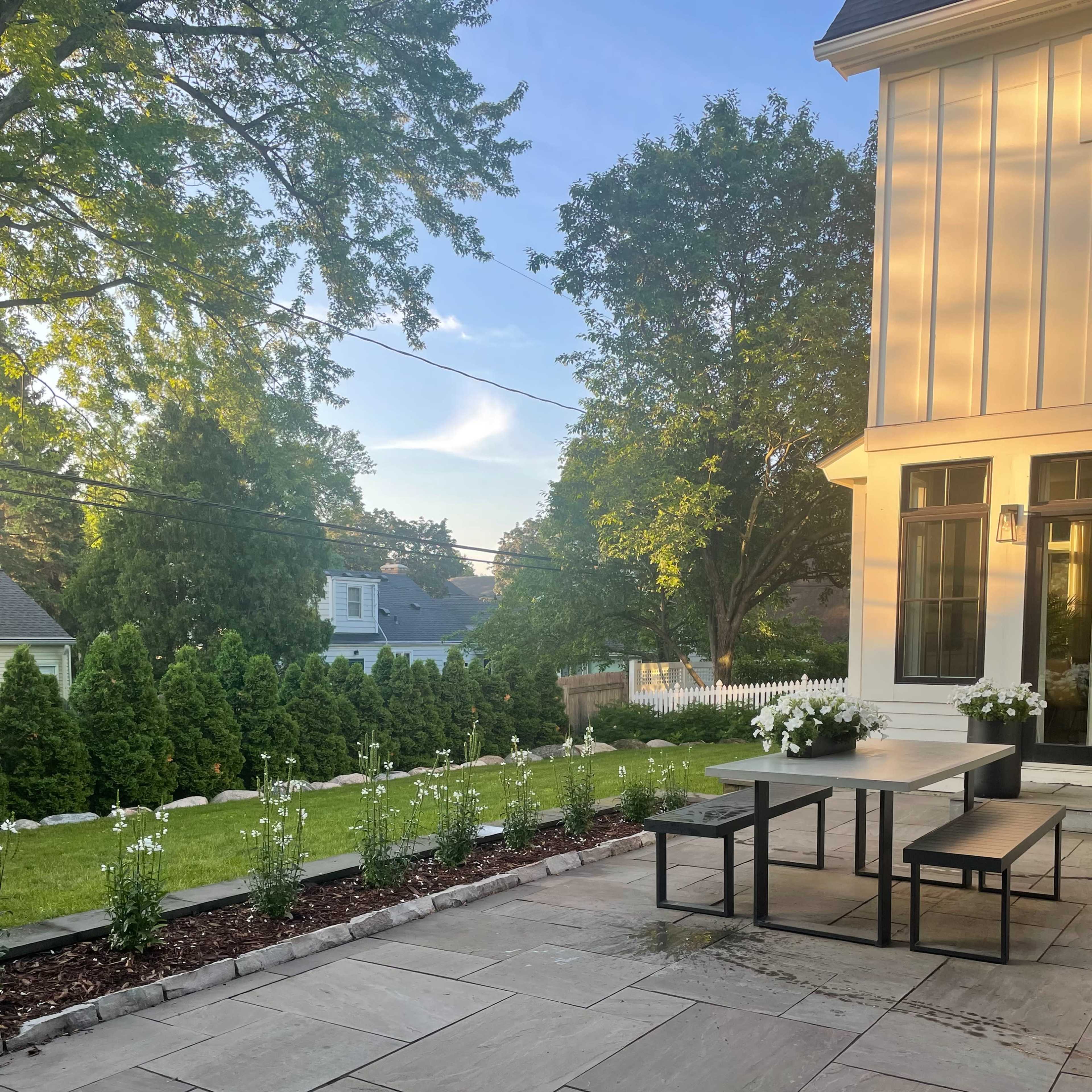A patio area with a stone surface, a table and bench set, and neatly arranged flower beds and shrubs in the background.