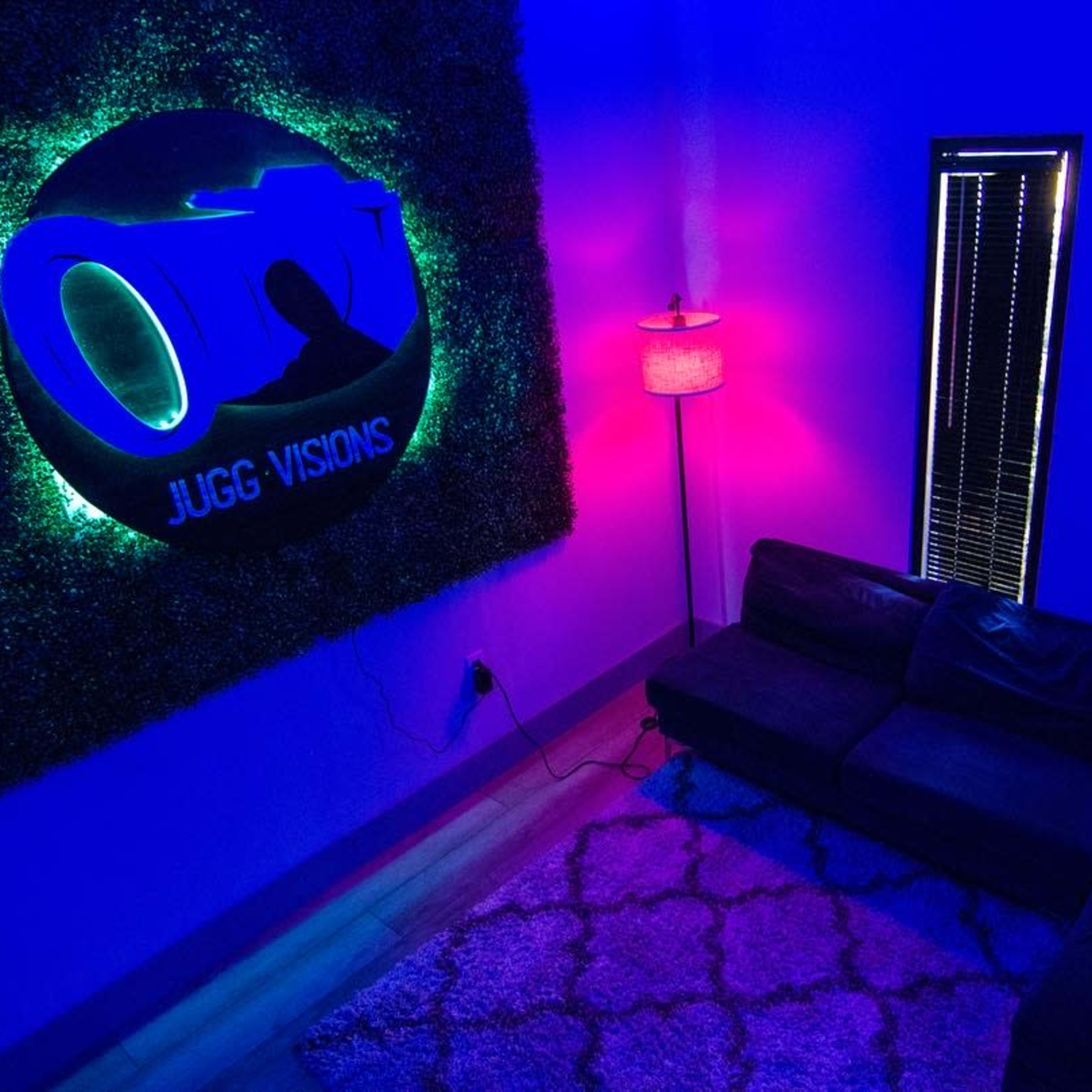 A dimly lit room with blue and purple lighting, featuring a neon sign that reads "JUGG VISIONS" on a textured wall, a lamp beside a dark sofa, and a window with blinds.