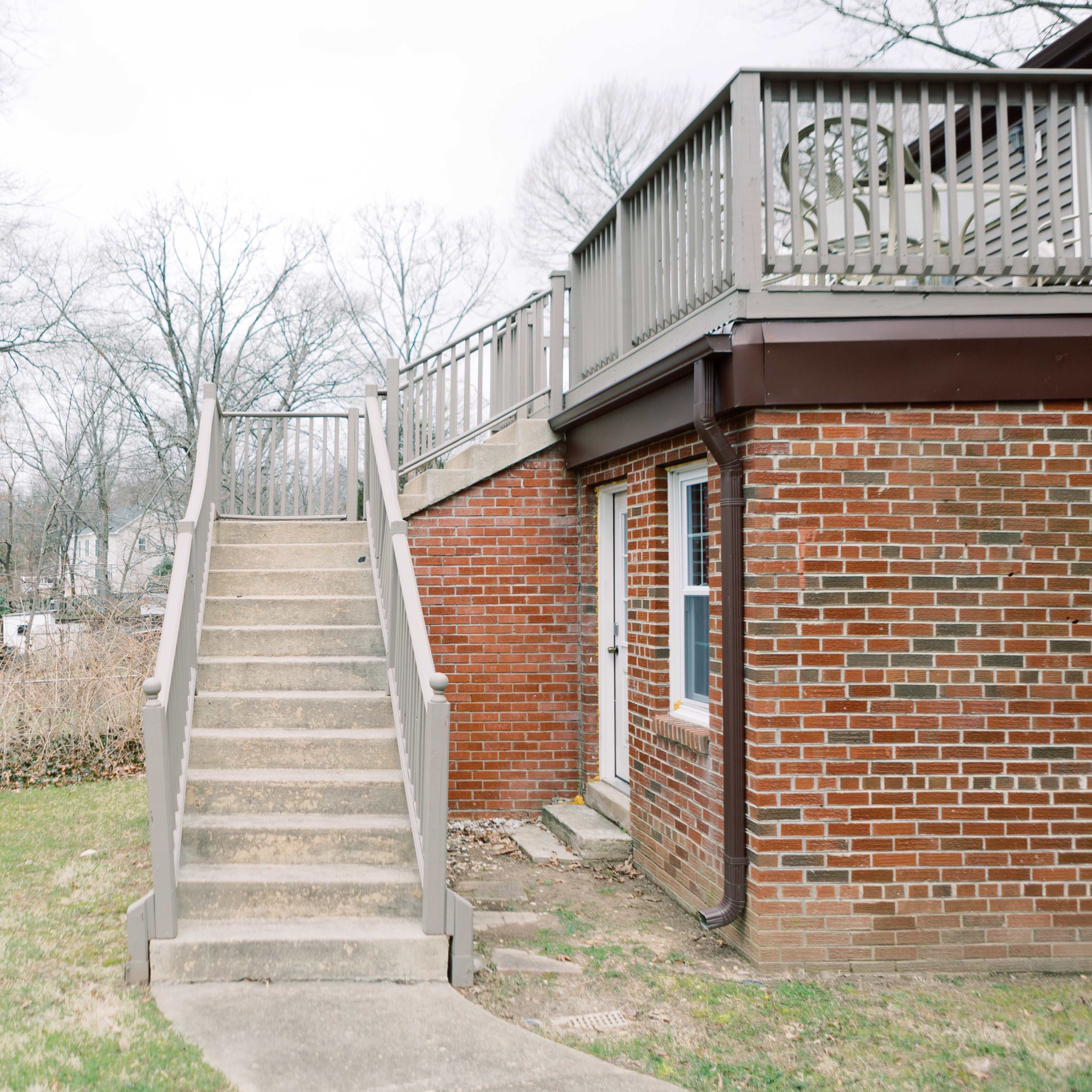 A set of concrete steps leads up to a wooden deck adjacent to a brick building in a treed yard.
