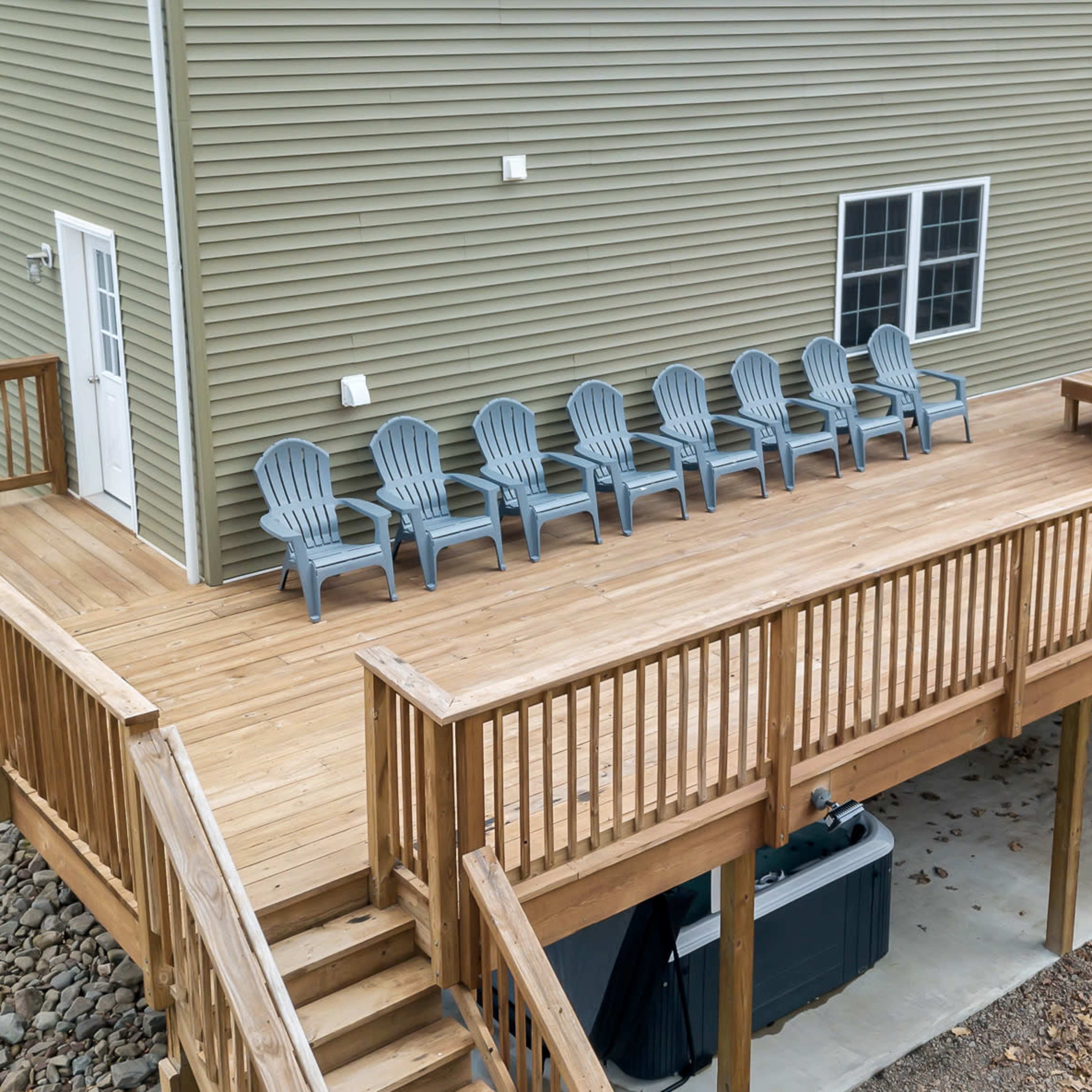 A wooden deck with several gray seated chairs arranged along the railing and a central table, adjacent to a green house.